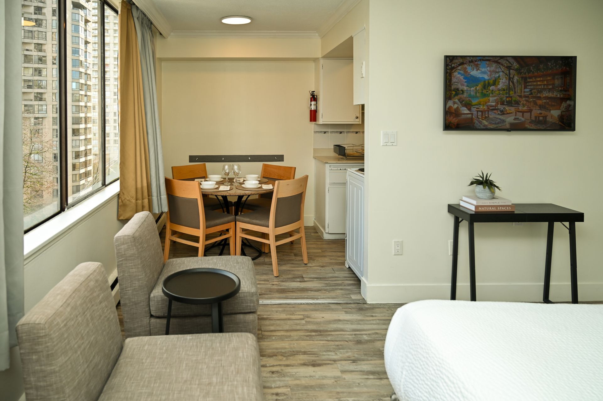 A brightly lit hotel room with a dining set, two gray chairs, a small side table, and a black console table with art.