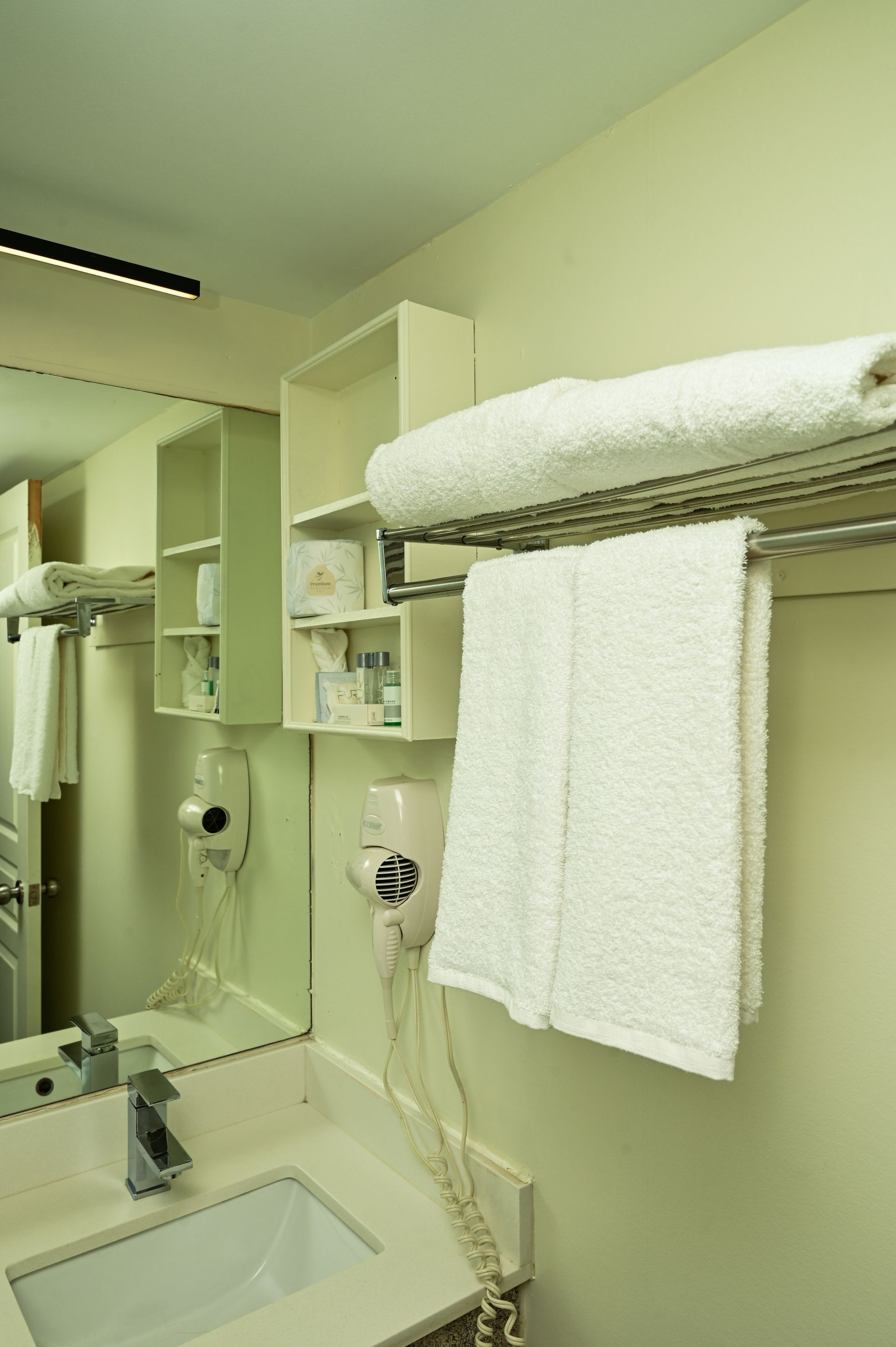 A hotel bathroom with a sink, mirror, wall-mounted hairdryer, shelving unit, and white towels on a metal rack.