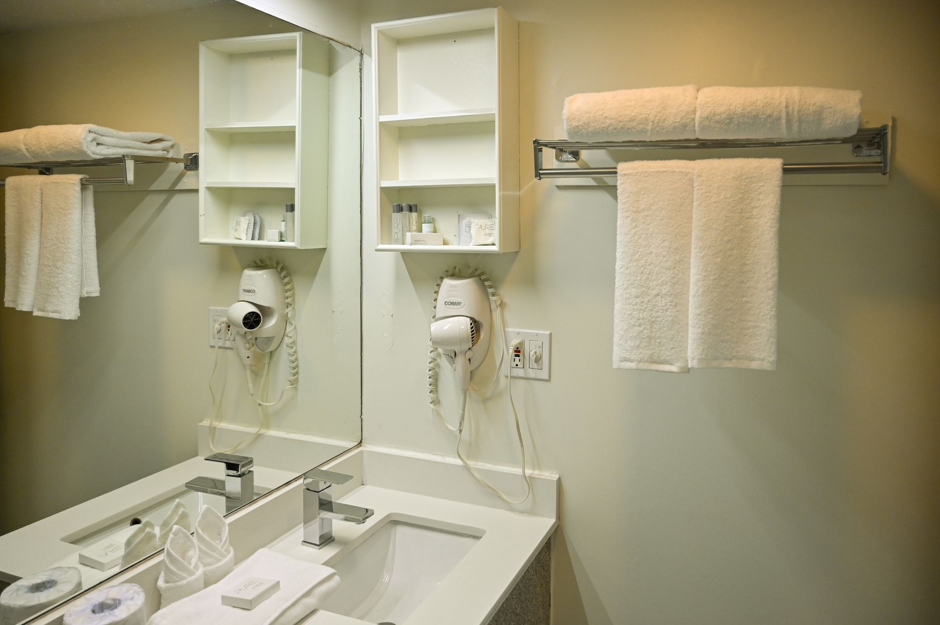 A clean hotel bathroom vanity with a mirror, shelves, hair dryer, and white towels hanging on a rack.
