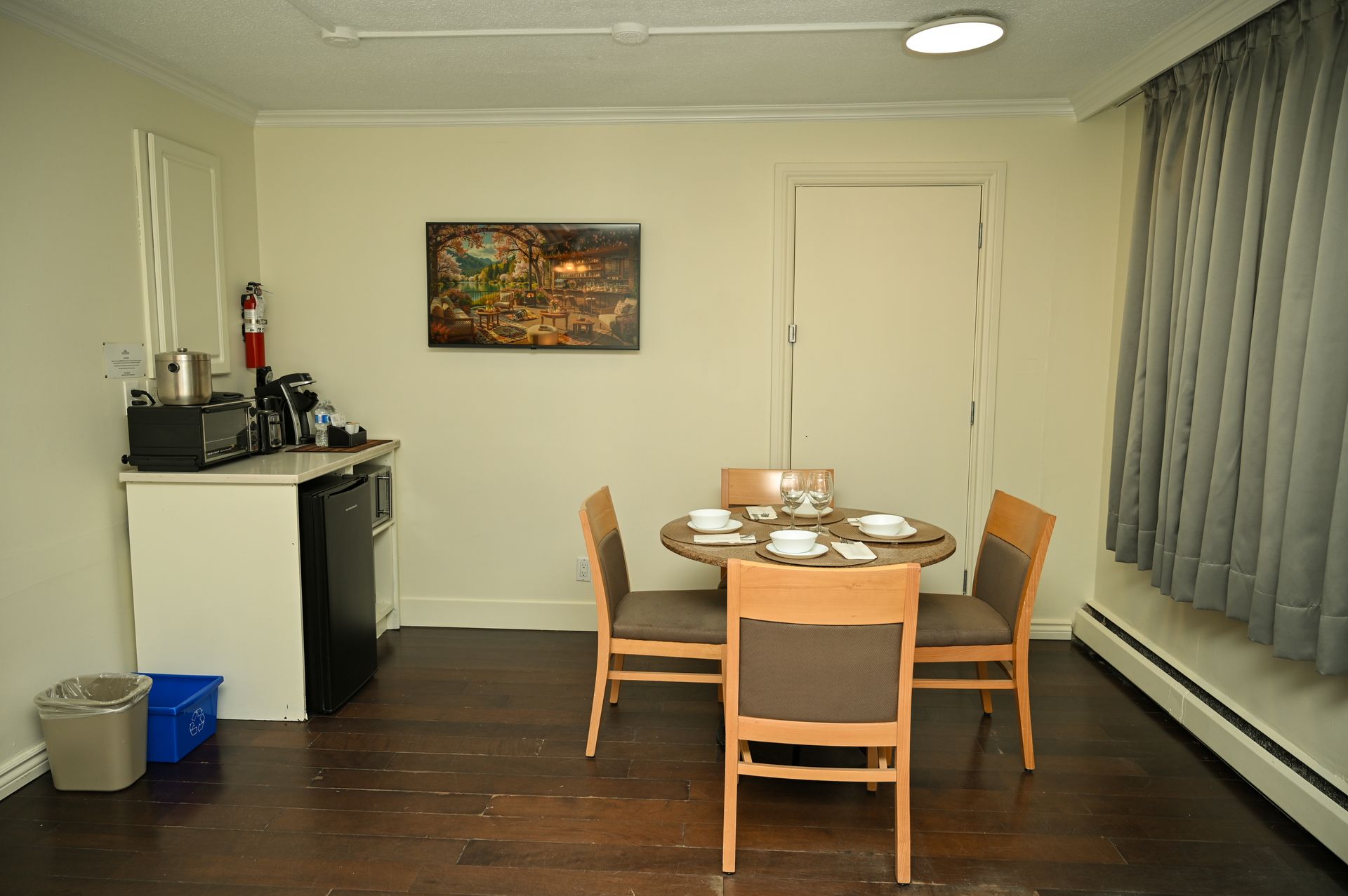 A dining area with a round wooden table, four chairs, a small kitchen counter with appliances, and a wall picture.