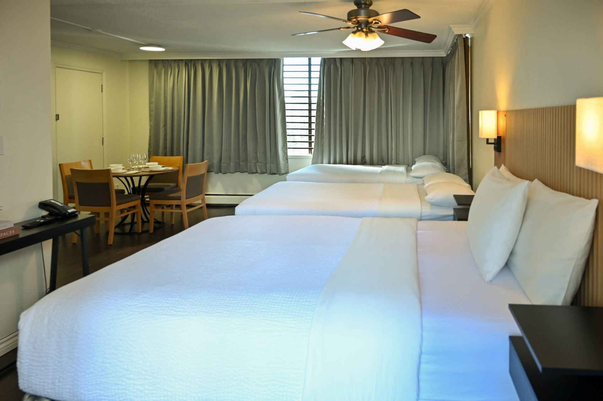 A hotel room with two white-made beds, a dining table with chairs, and curtains covering a window.