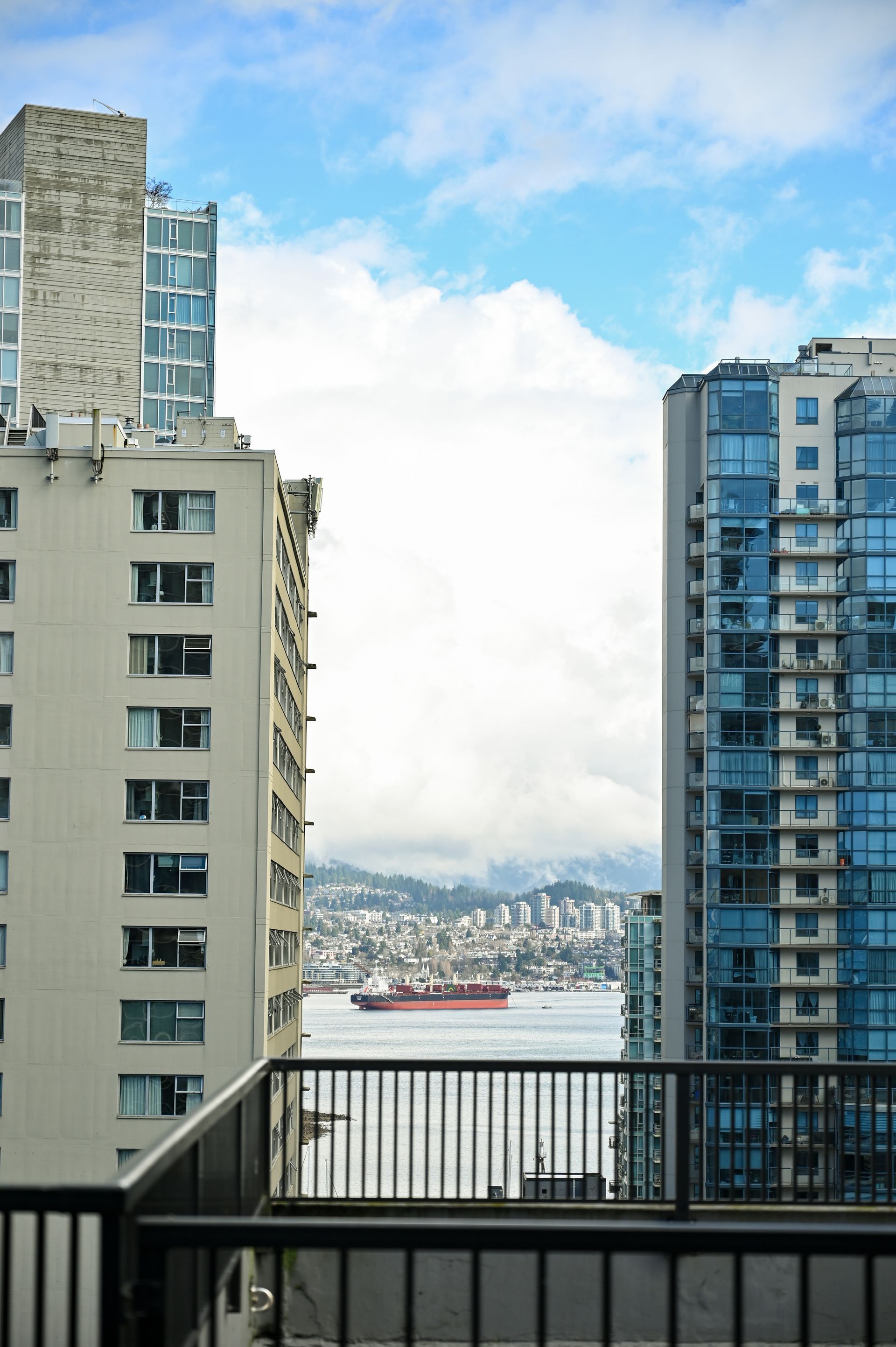 A view of a harbor with a ship between two tall apartment buildings, framed by a balcony railing under a cloudy sky.