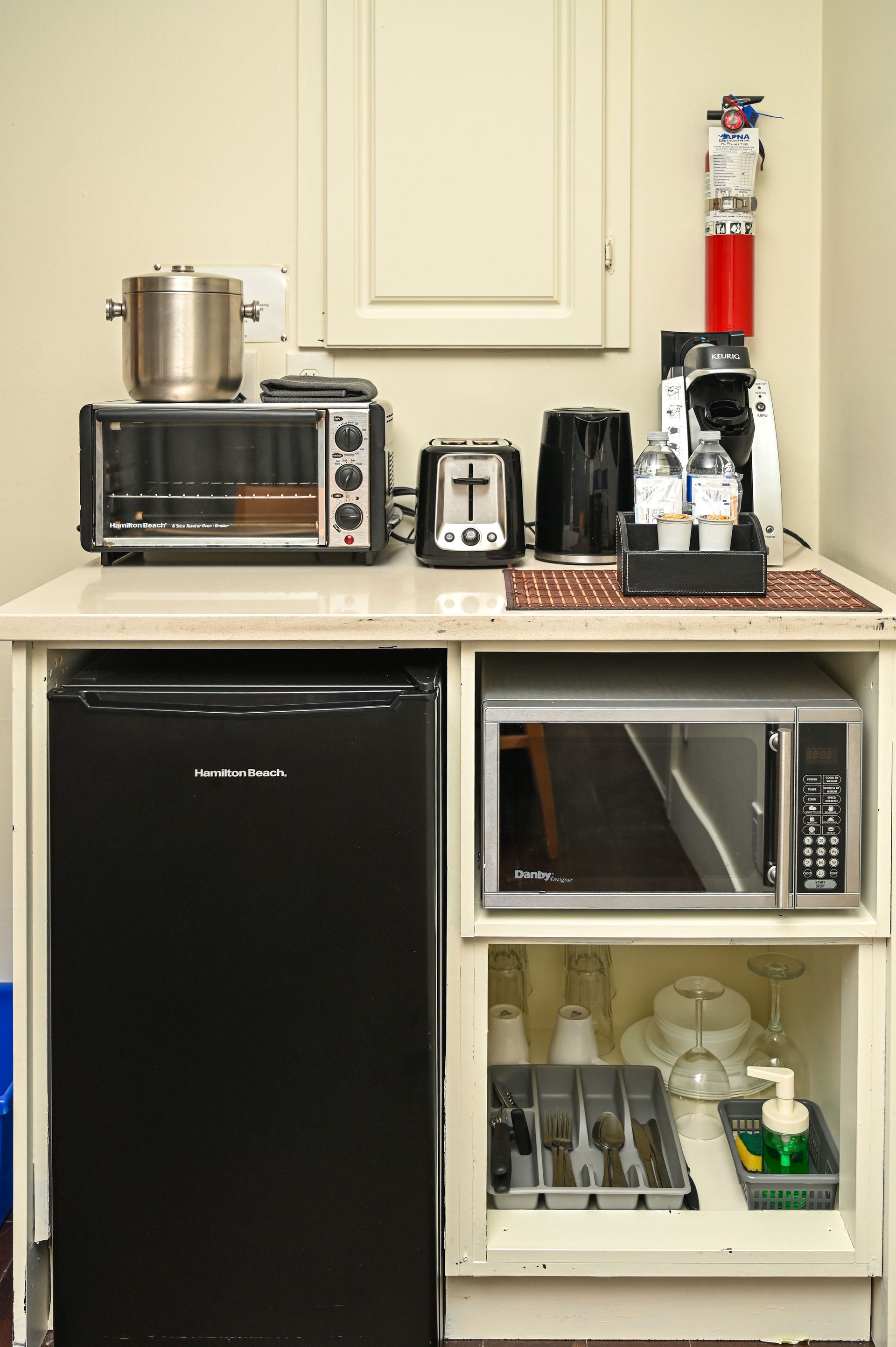 A kitchenette with a black mini-fridge, microwave, toaster, coffee maker, and shelf with kitchen supplies.