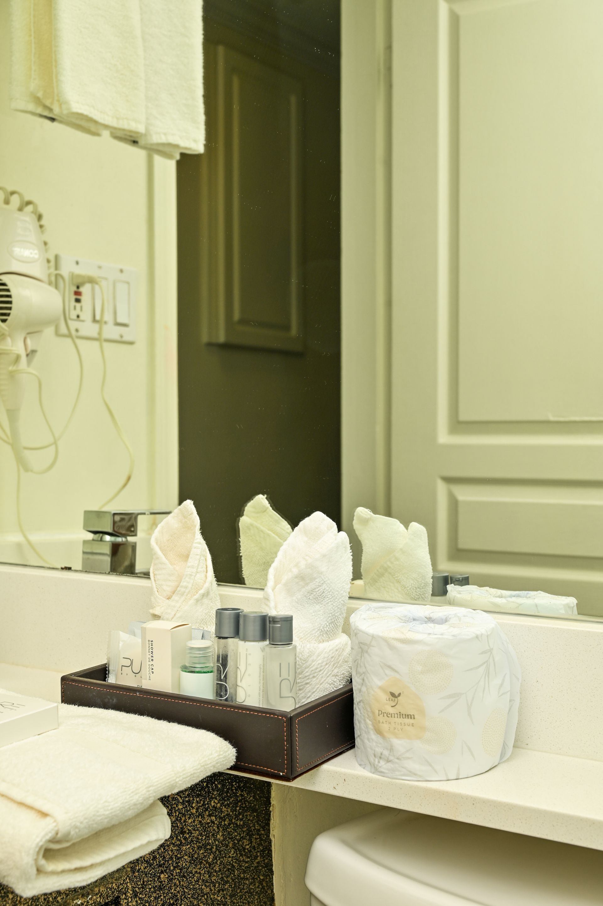 Hotel bathroom vanity with rolled white towels, toiletries in a brown tray, and a roll of toilet paper on the counter.