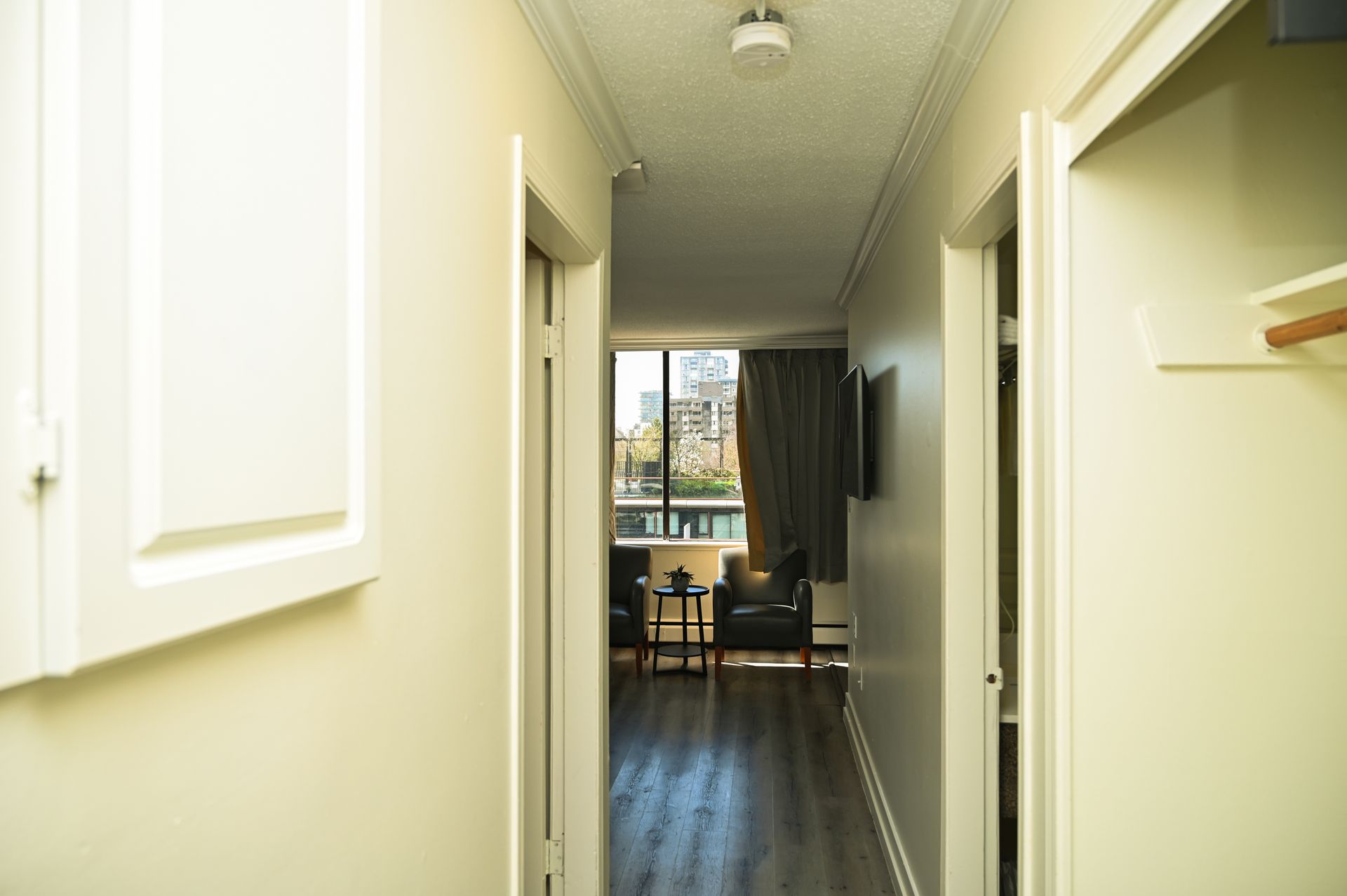A brightly lit hallway leading to a view of city buildings through a window with dark curtains and two chairs.