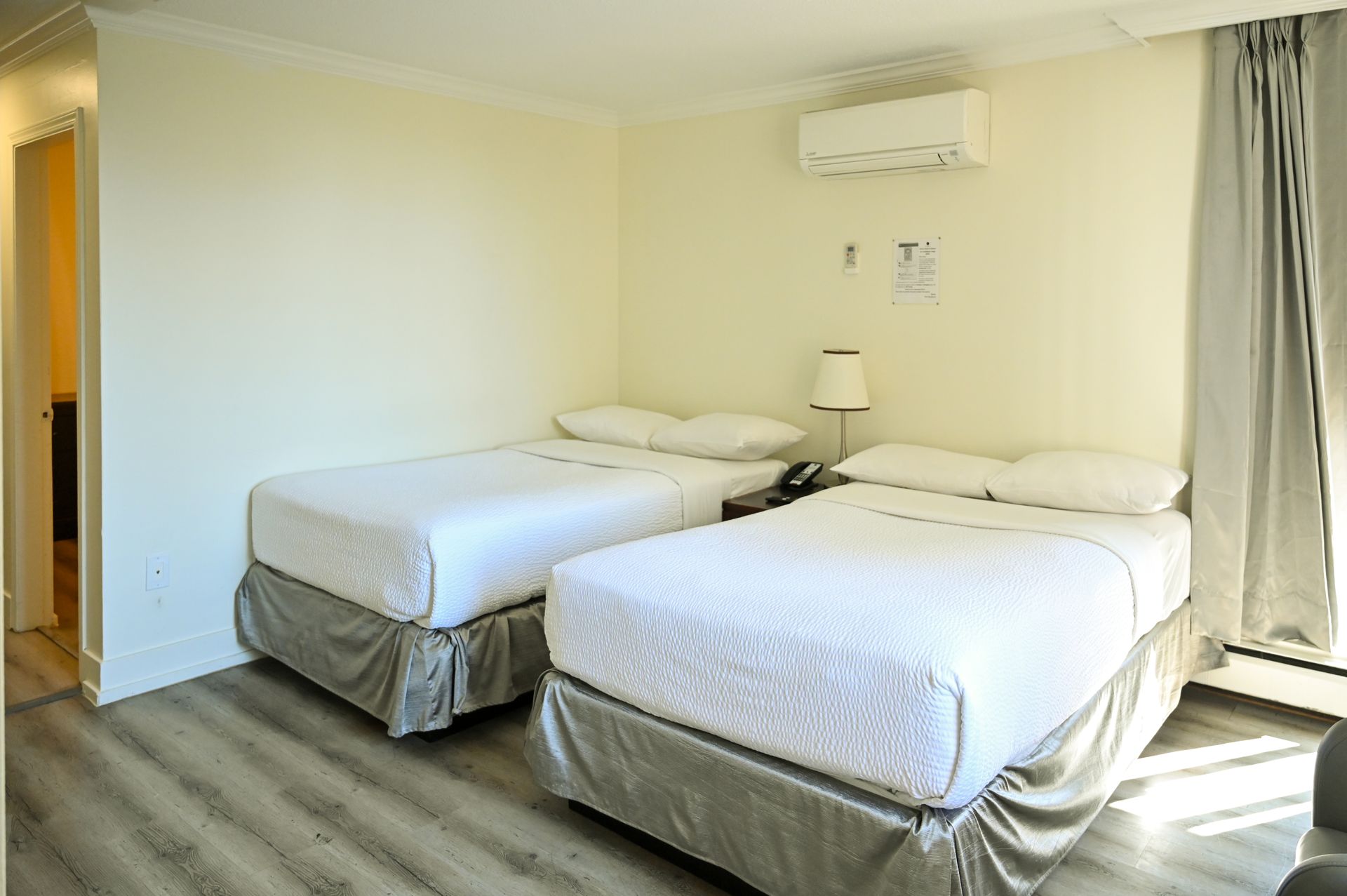 A hotel room with two beds with white bedding, gray skirts, light walls, light wood flooring, and curtains.
