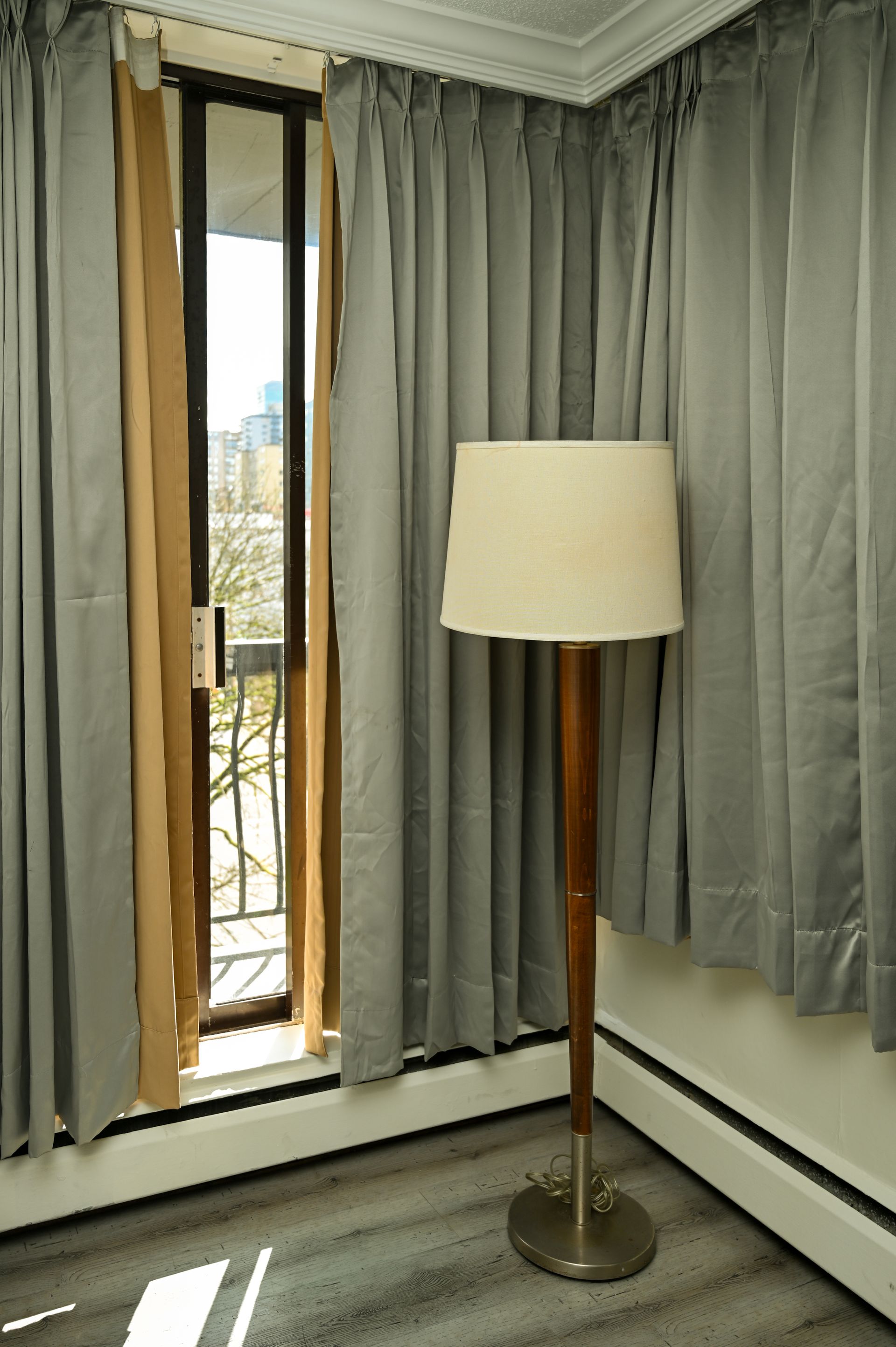 A wooden floor lamp stands in a corner next to floor-to-ceiling grey curtains and a partially open balcony door.
