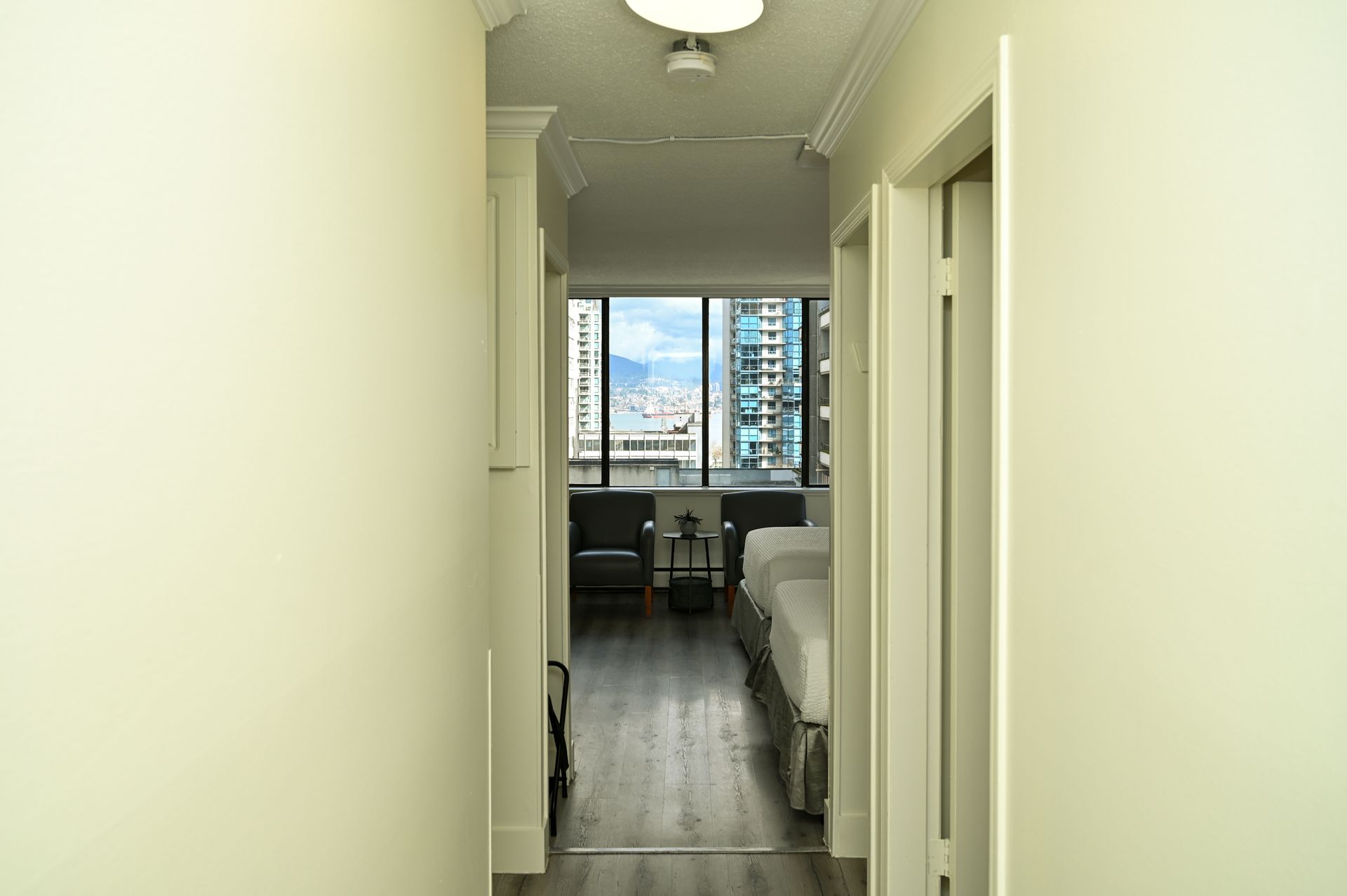 A hallway with light walls leads toward a windowed room with two beds and dark chairs overlooking a city view.