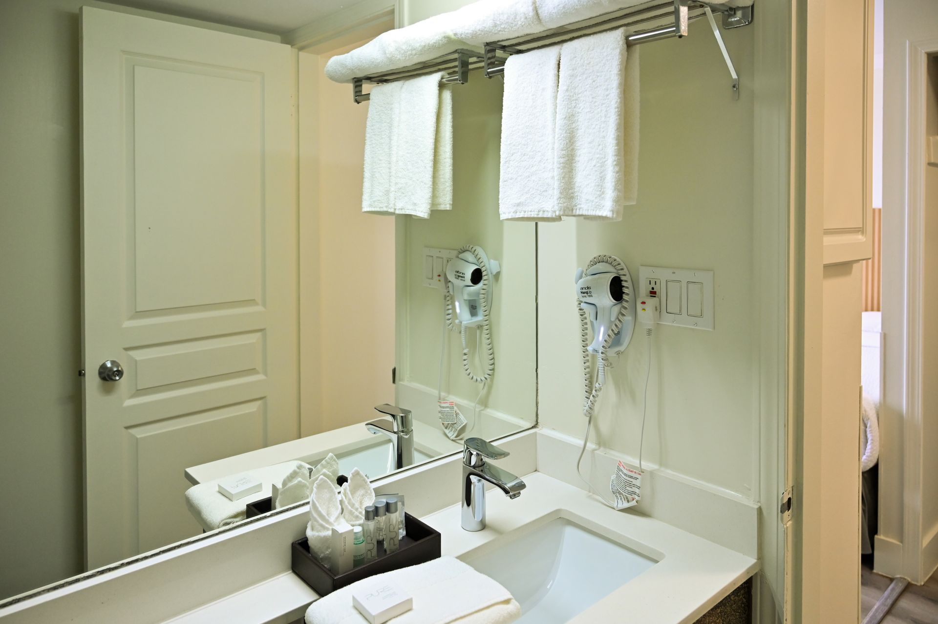 A hotel bathroom vanity with a sink, mirror, wall-mounted hairdryer, white towels on a rack, and toiletries.