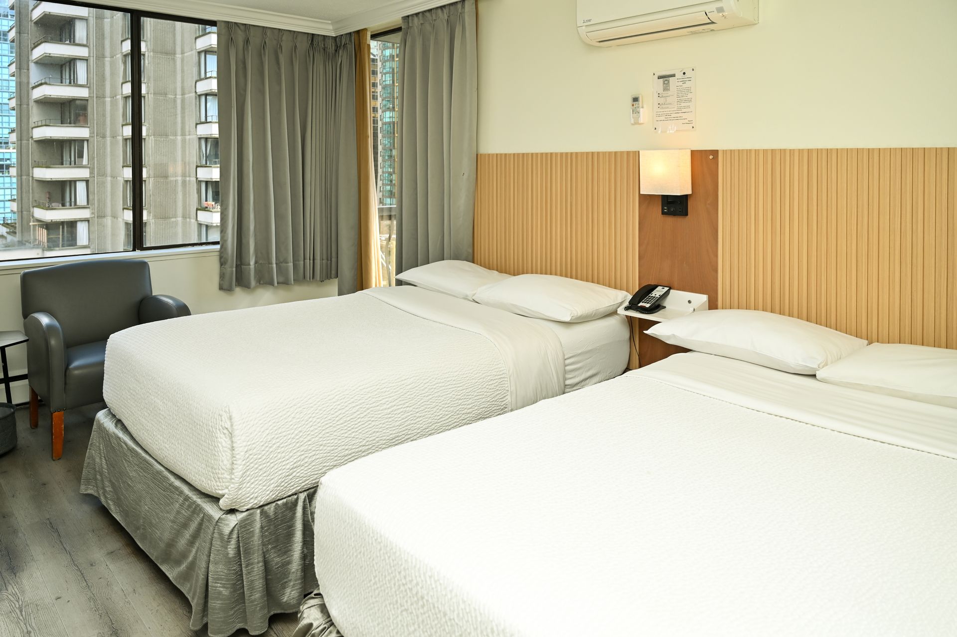 A hotel room with two white-bedded twin beds, a grey armchair, light wood headboards, and curtains over a city window.