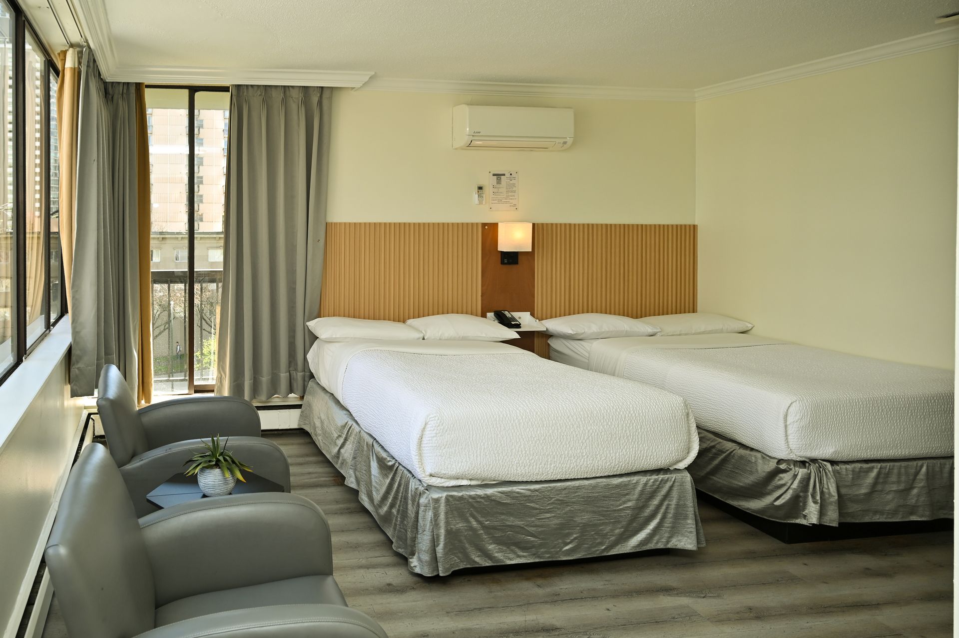 A hotel room with two white beds, a wooden headboard, two gray armchairs, and floor-to-ceiling windows with gray curtains.