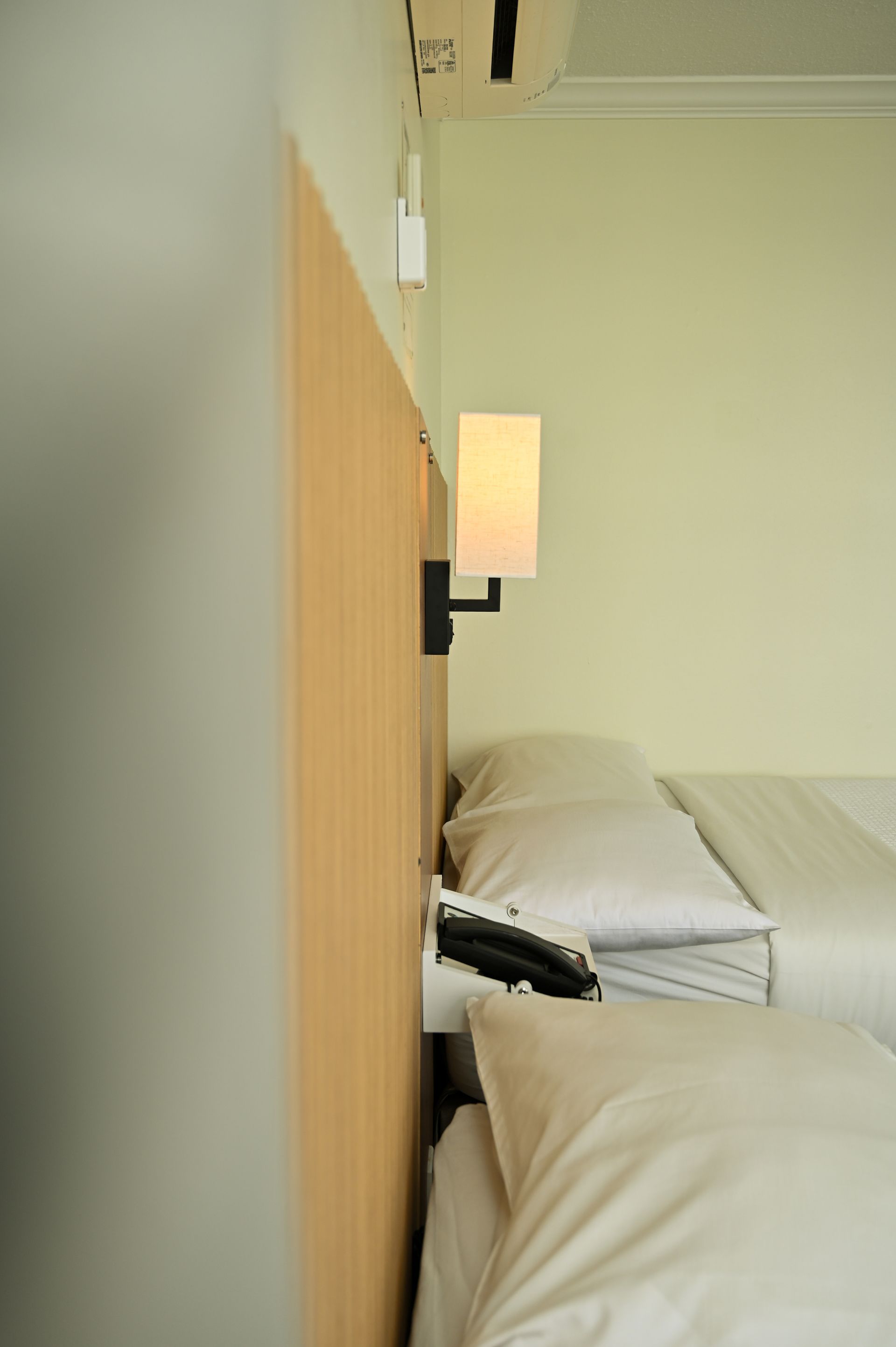 A side view of a hotel room with white beds, a wall-mounted reading light, and a telephone on a wooden headboard.