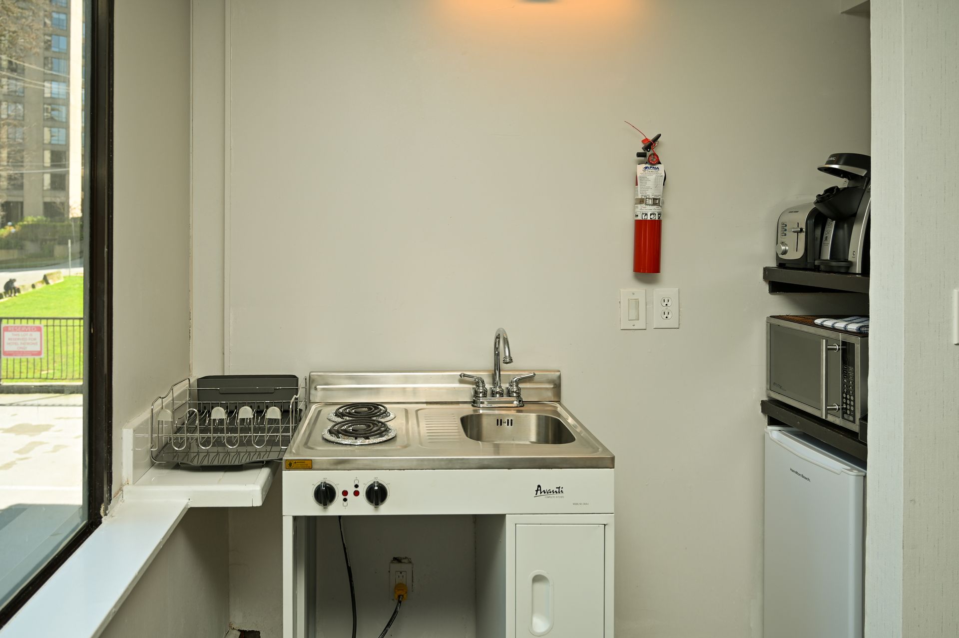 A compact kitchenette with a stovetop, sink, dish rack, microwave, small refrigerator, and fire extinguisher.