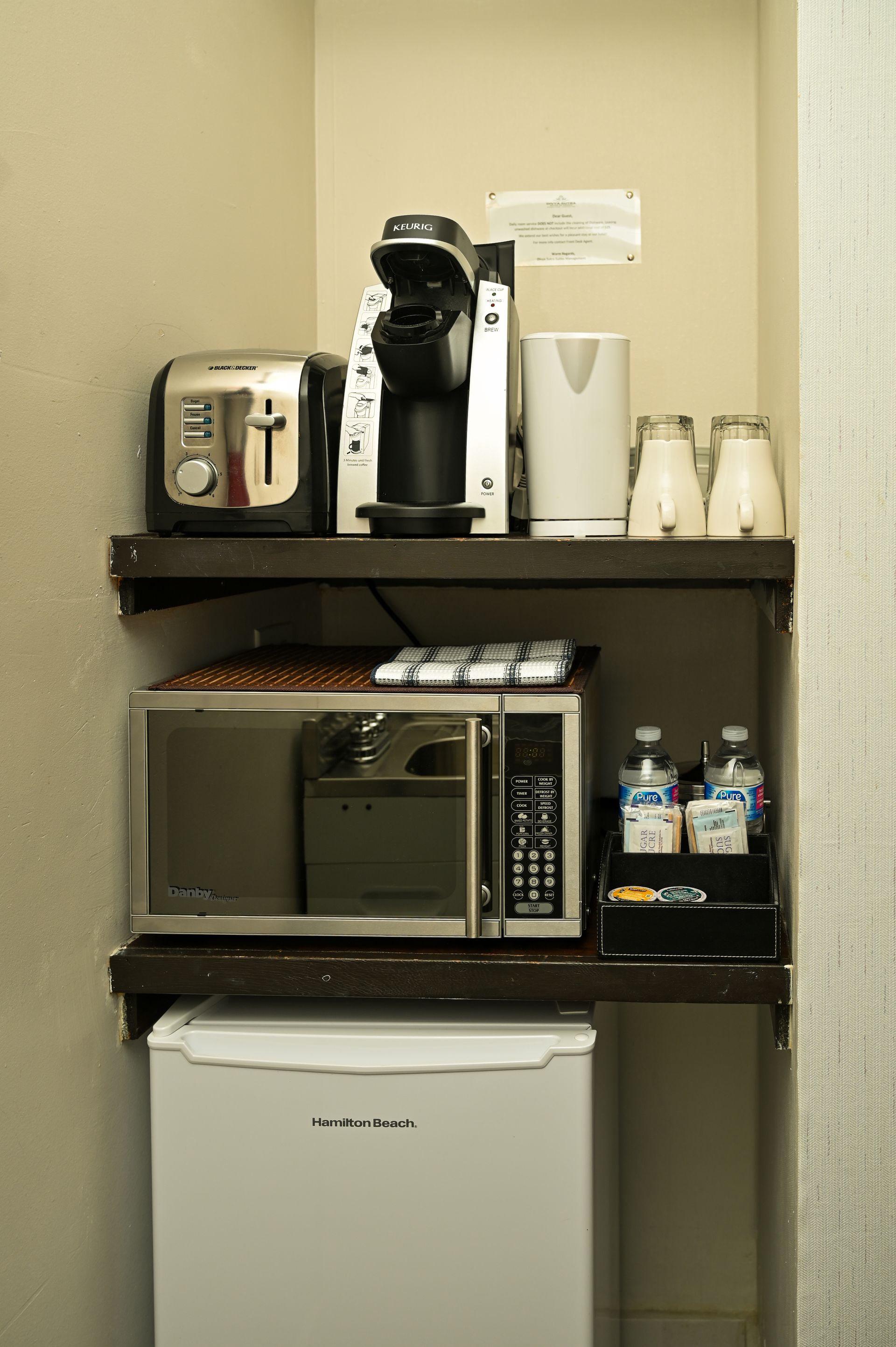 A hotel kitchenette nook with a toaster, coffee maker, kettle, microwave, mini-fridge, and water bottles on white shelves.
