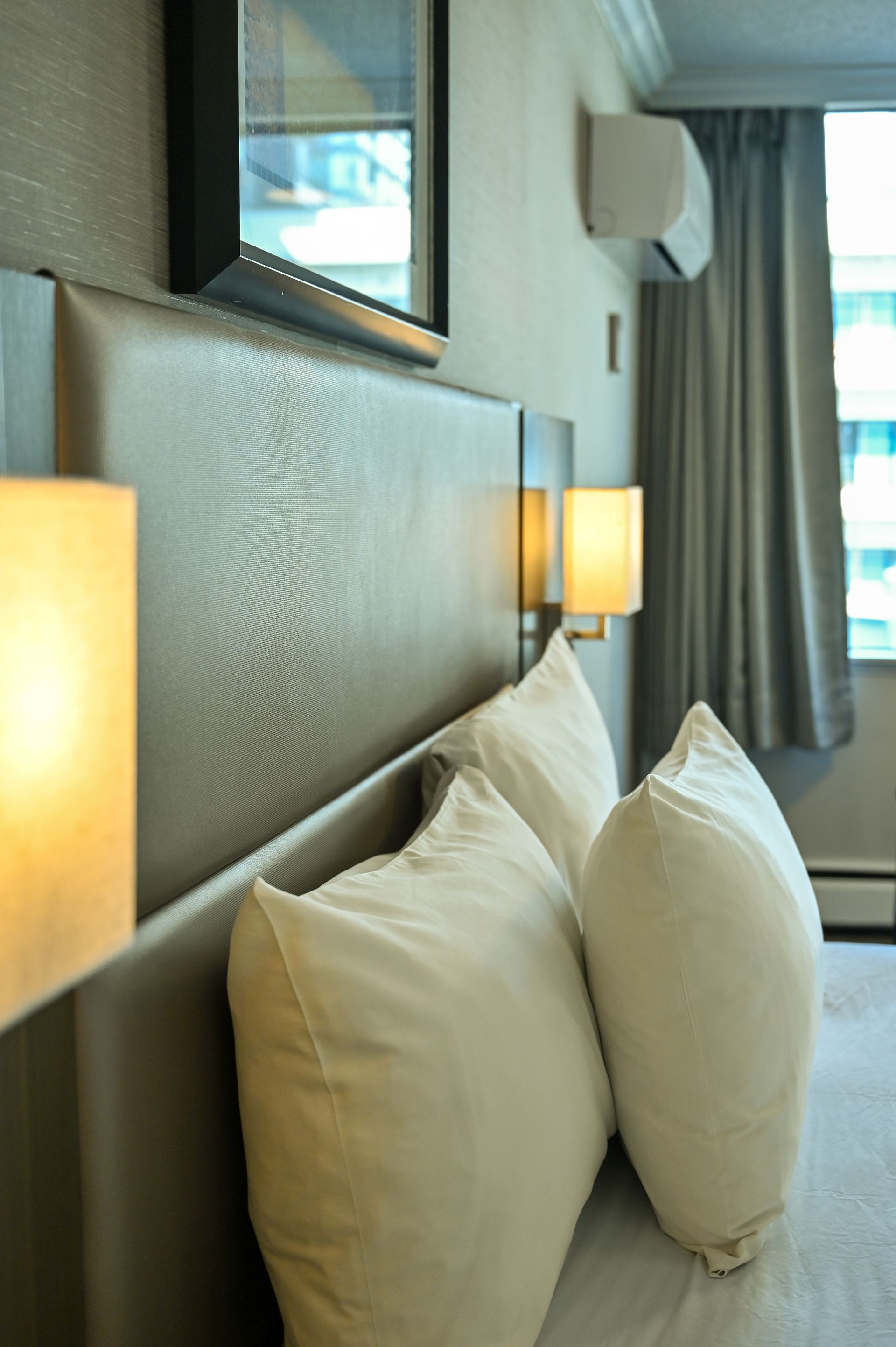 A close-up of a modern hotel bed headboard with textured beige upholstery, two white pillows, and warm bedside lamps.