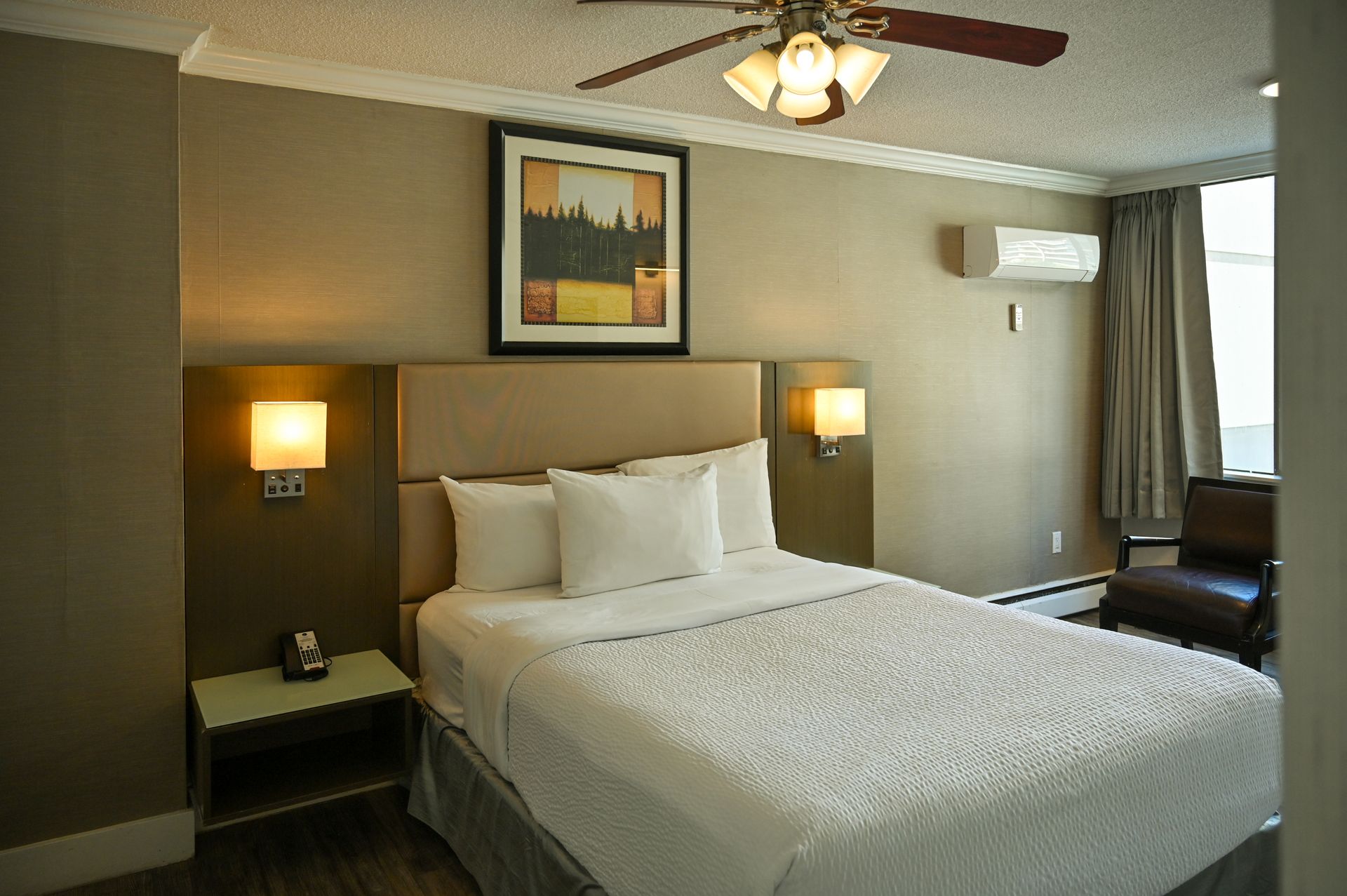 A hotel room with a queen bed, two bedside lamps, a dark armchair, and a wall-mounted air conditioner.
