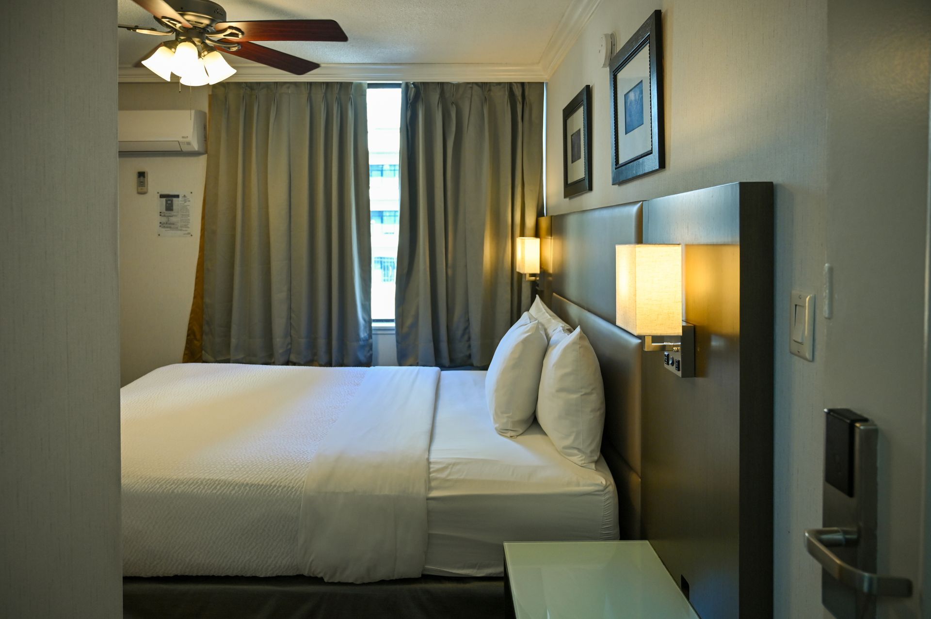 A hotel room featuring a bed with white linens, grey curtains, a ceiling fan, and wall-mounted lights above the headboard.