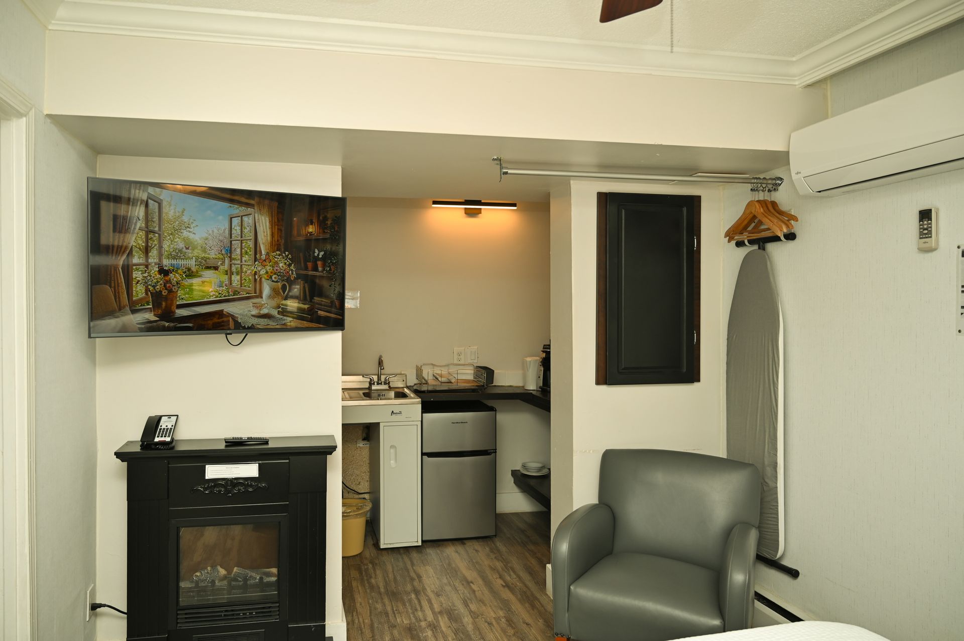 A hotel room corner featuring a wall-mounted TV, black electric fireplace, mini-fridge, grey armchair, and ironing board.