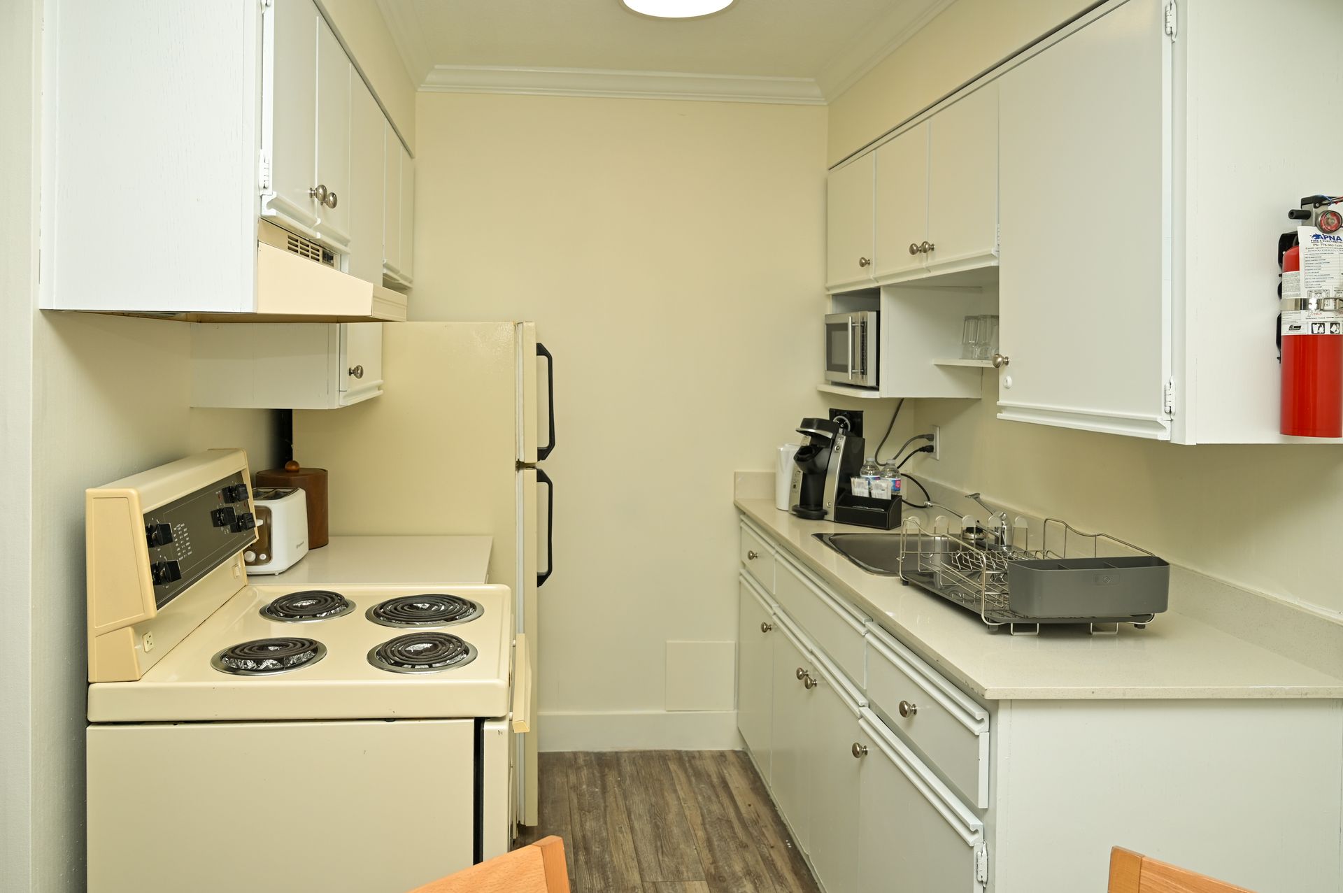 A small, white kitchen featuring an electric stove, refrigerator, cabinets, countertops, and a mounted fire extinguisher.