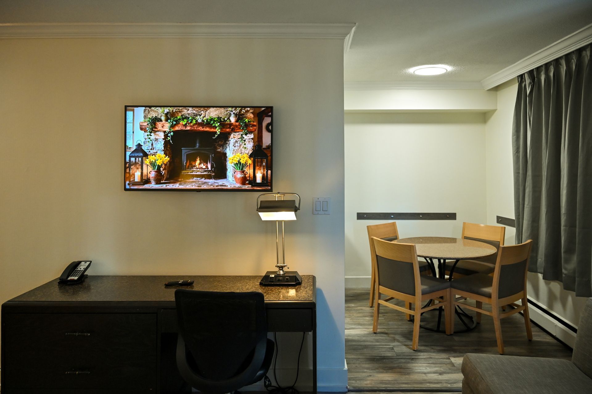 A desk with a computer monitor and lamp, next to a dining area with a round table and chairs in a neutrally toned room.