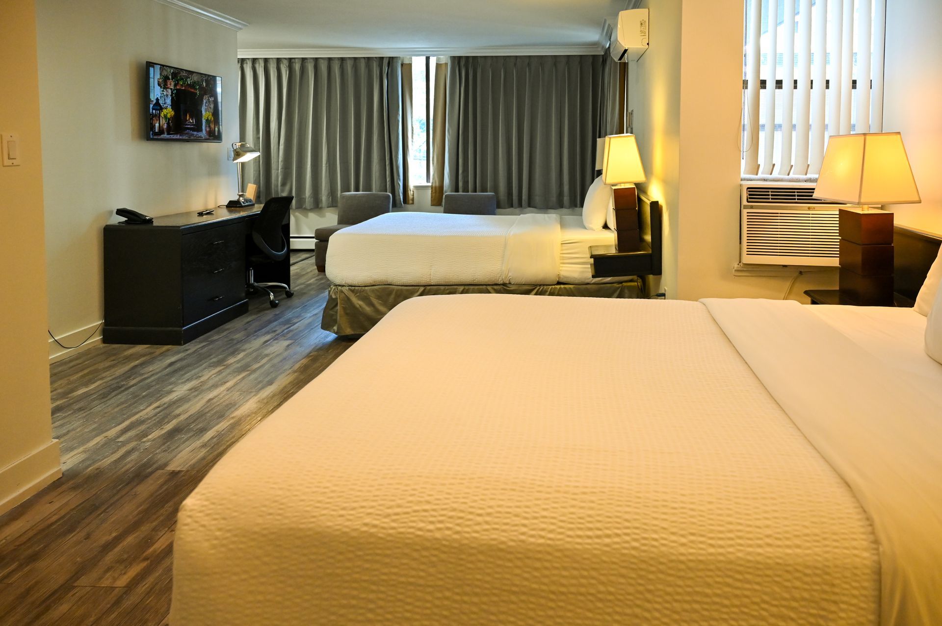 A hotel room with two white beds, a dark desk with a chair, gray curtains, and a lamp on a bedside table.