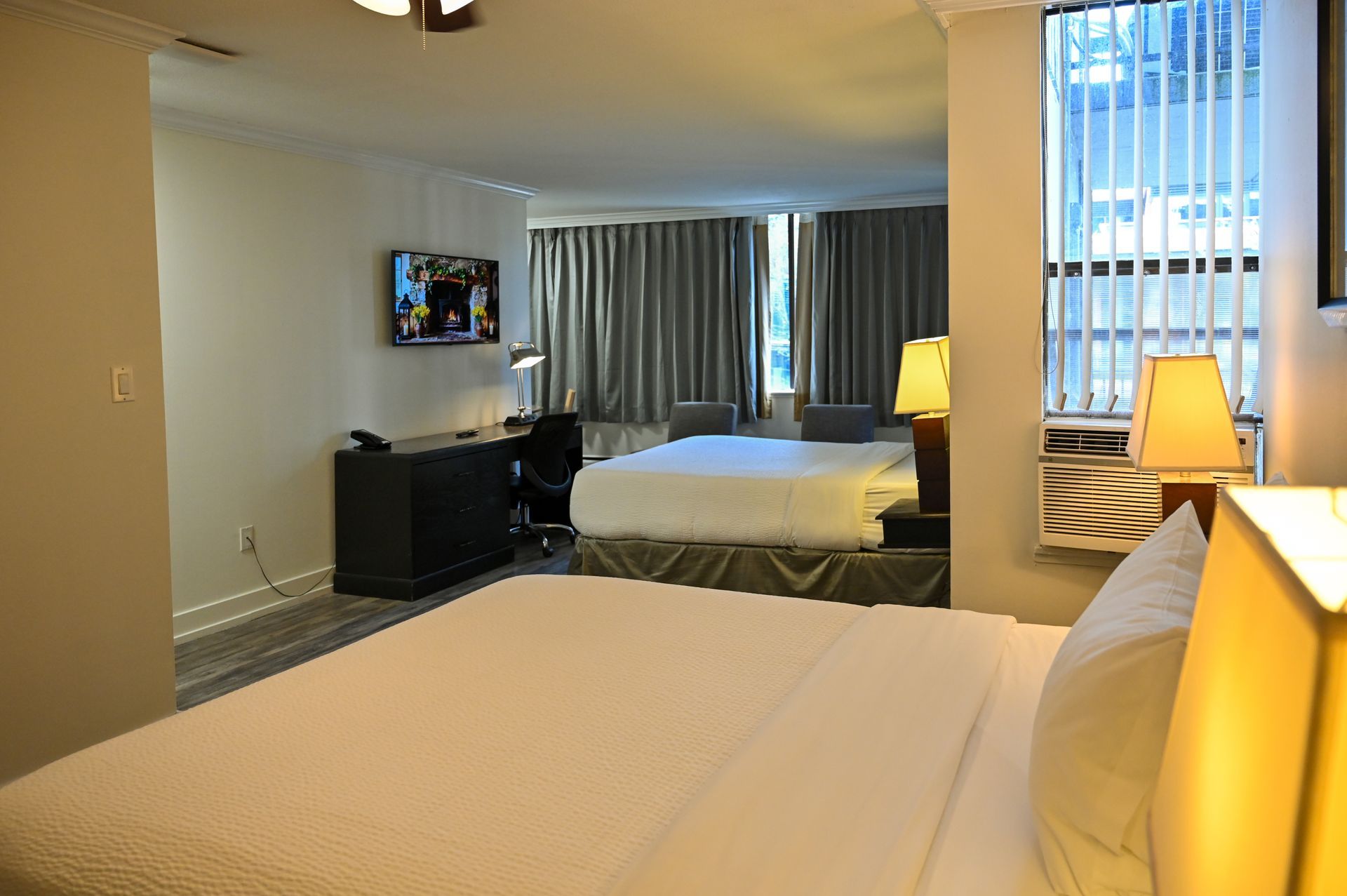 A hotel room featuring two beds, a desk area with a television, and a window with blinds.