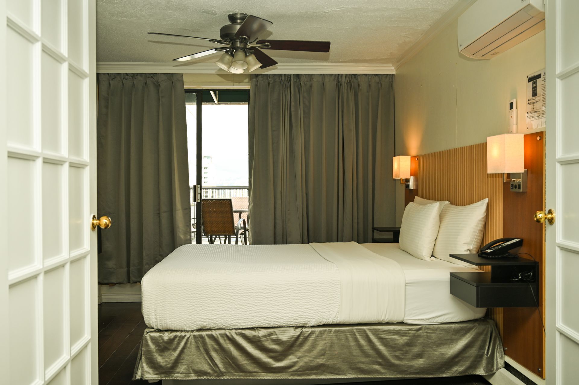A view through white French doors into a hotel room with a white bed, neutral curtains, and wall-mounted bedside lamps.