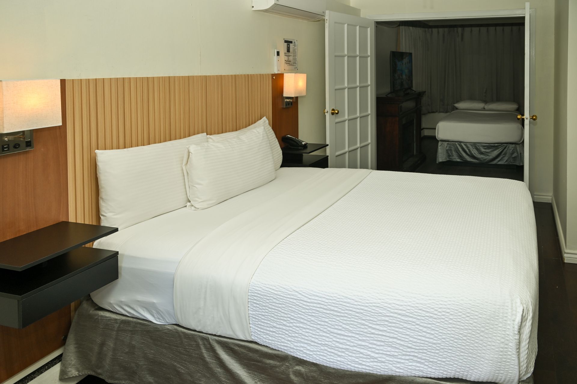 A hotel room featuring a bed with white linens and a light wood headboard, with an adjacent room visible through a door.