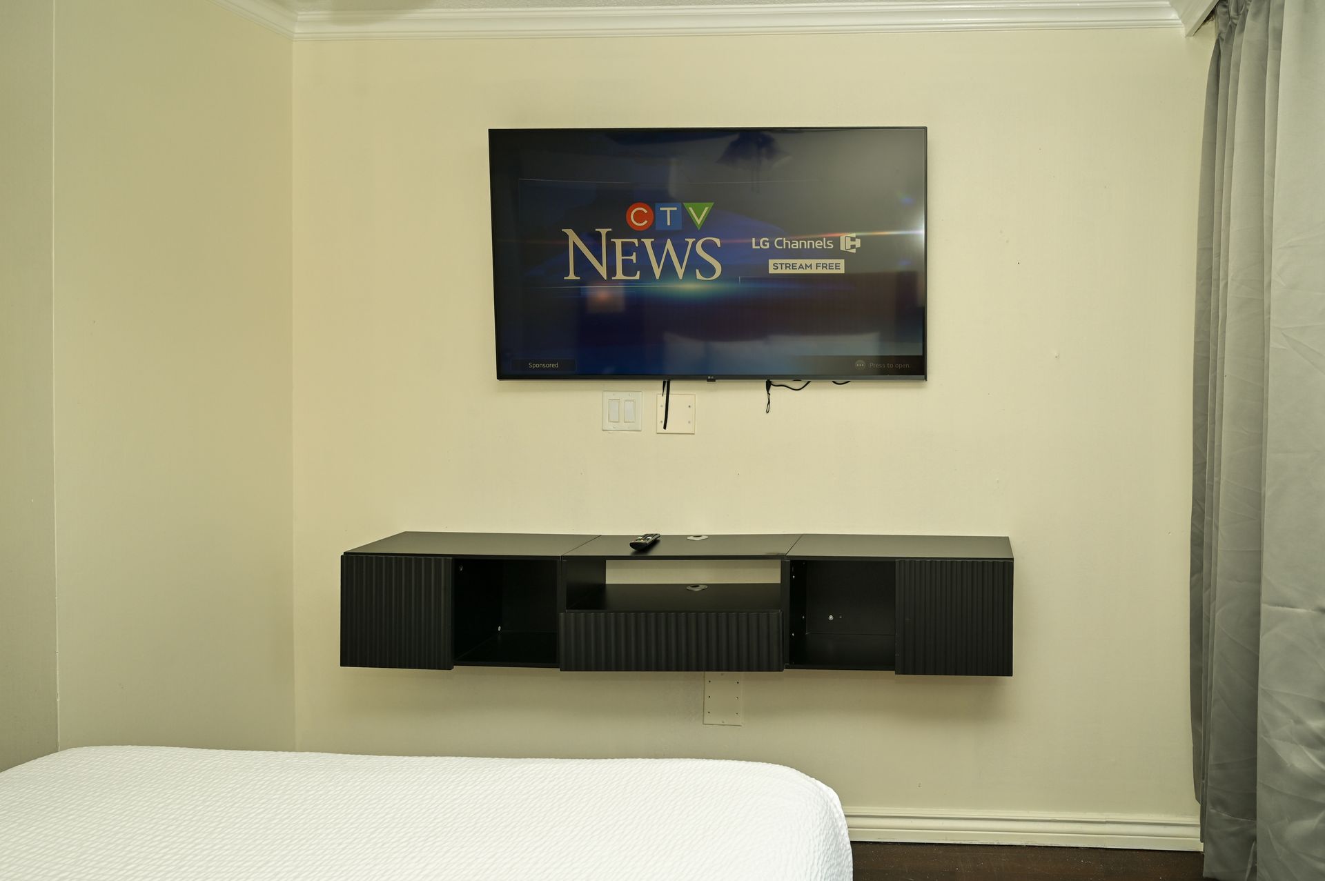 A wall-mounted TV showing CTV News hangs above a dark, floating shelf in a neutrally painted room with grey curtains.