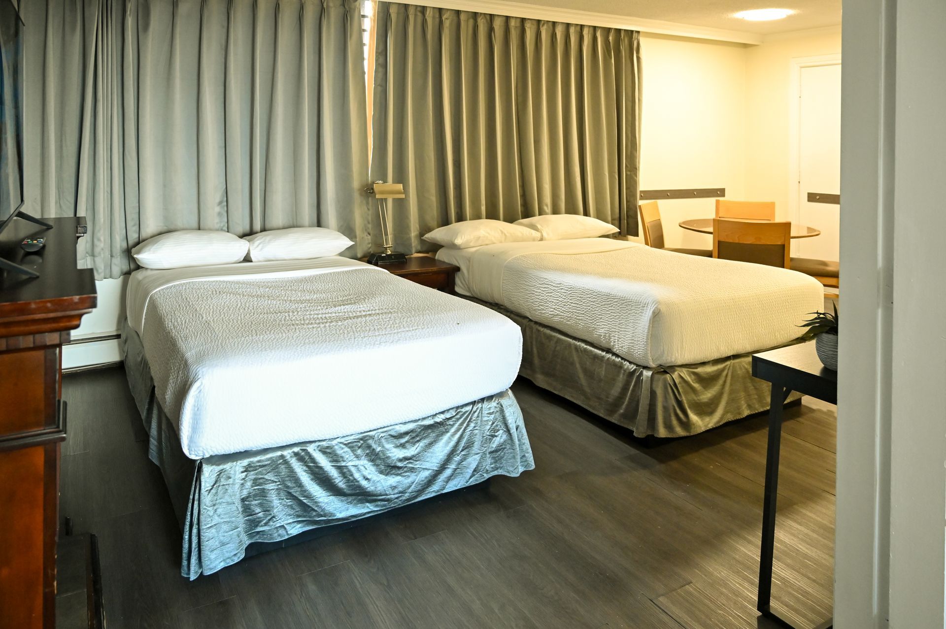 A hotel room with two white-bedded full beds, light grey curtains, and dark wood-look floors.