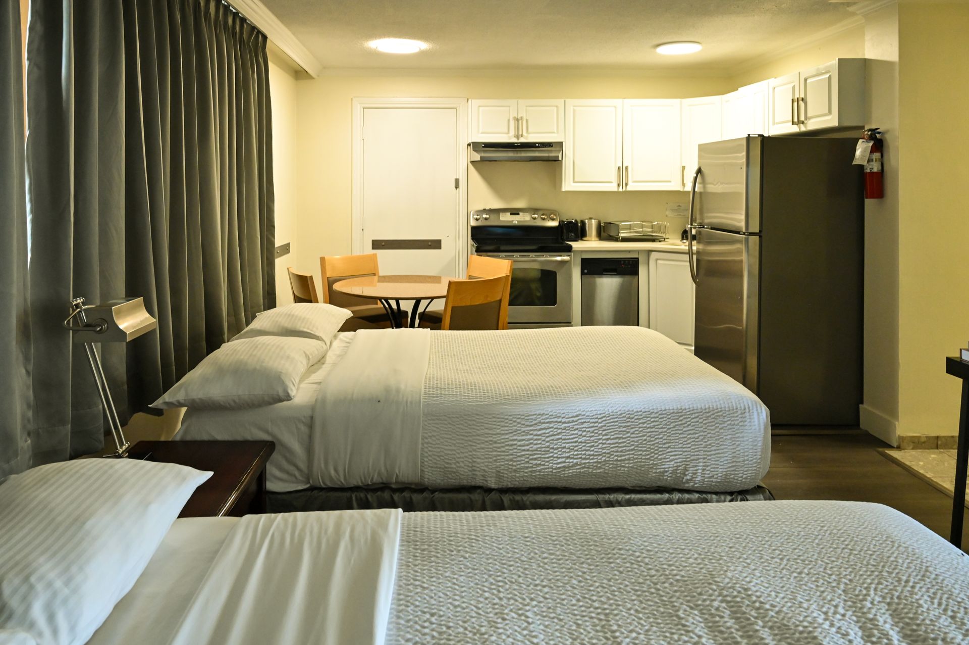 A hotel room featuring two beds in the foreground, a small dining table, and a kitchenette with appliances in the back.