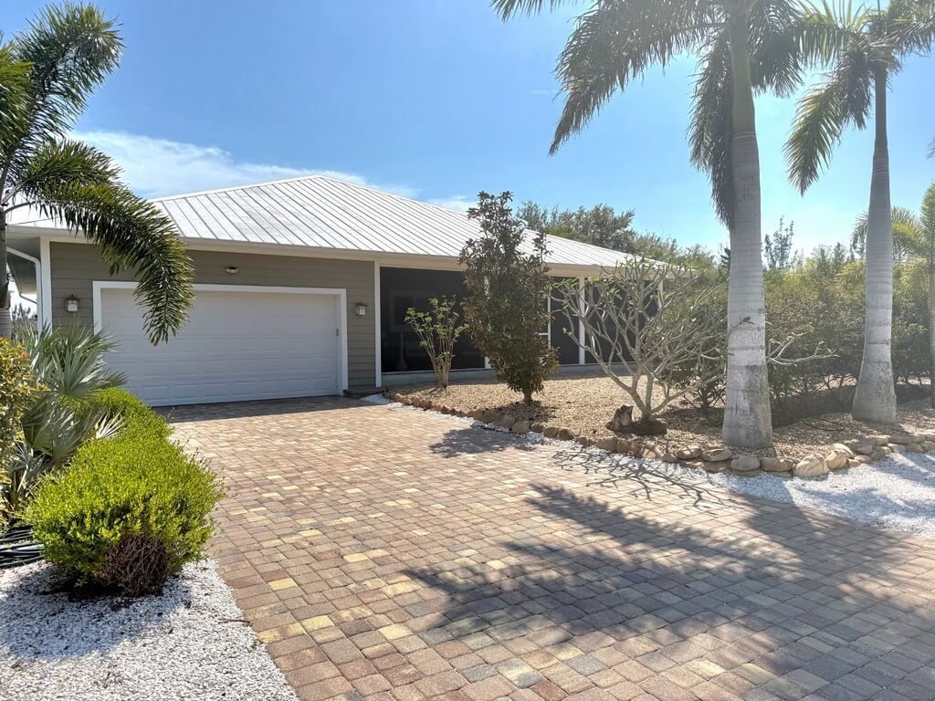 A tan single-story house with a white roof and garage, fronted by a brick driveway, lush bushes, and tall palm trees.