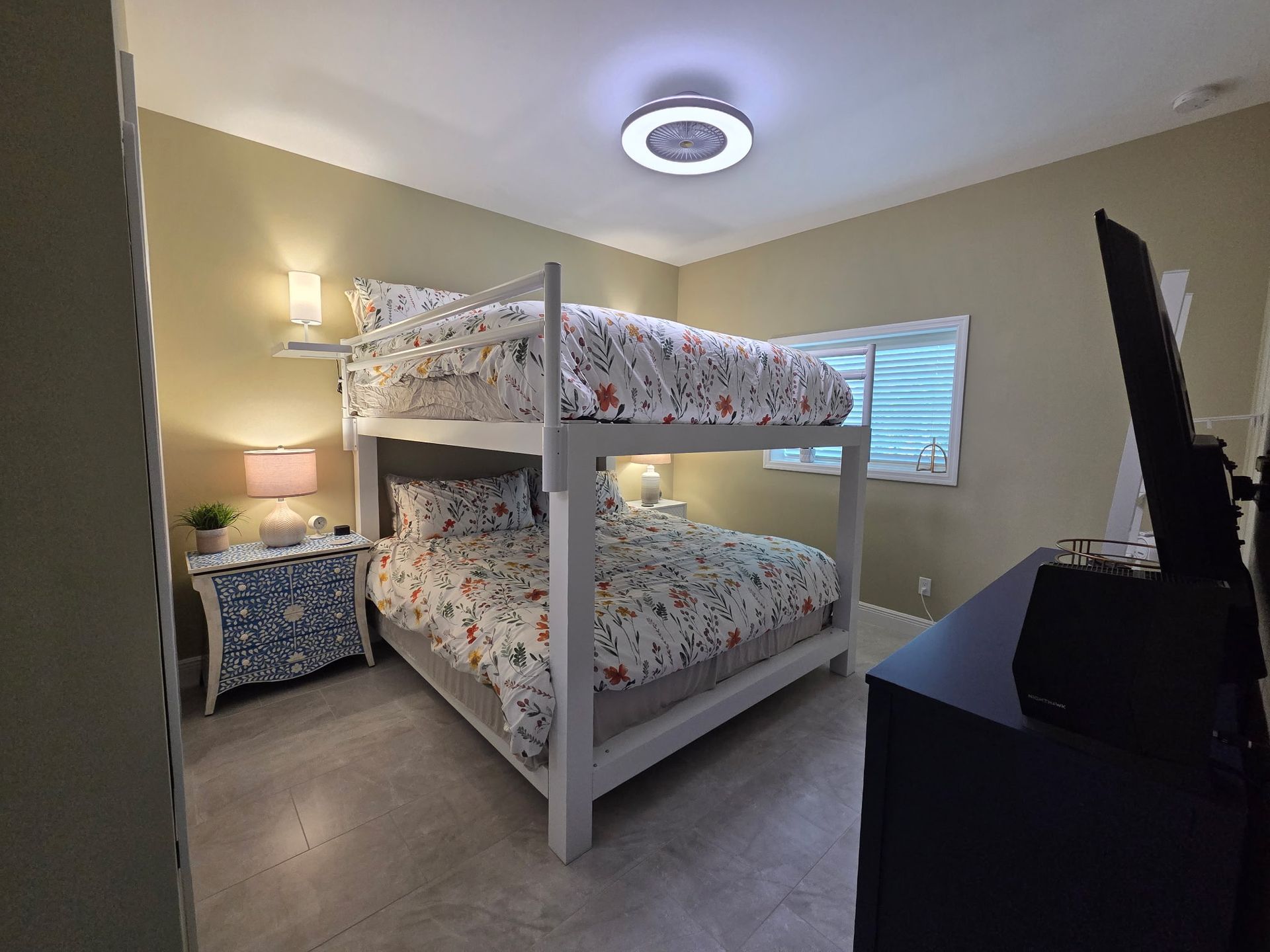 A bedroom with a white bunk bed featuring floral bedding, a small blue nightstand, and a black dresser with a TV.