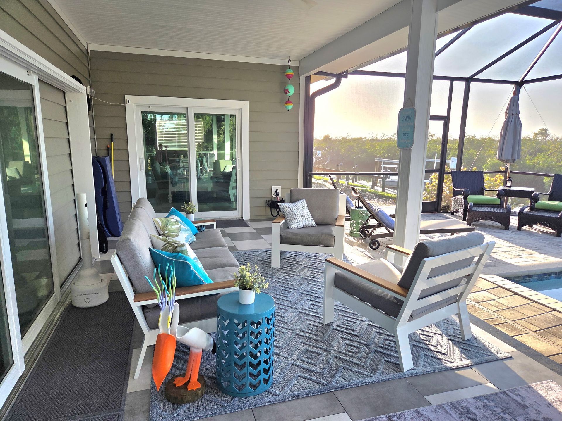 A covered patio with light wood furniture, blue accent pillows, a patterned area rug, and a pool in the background.