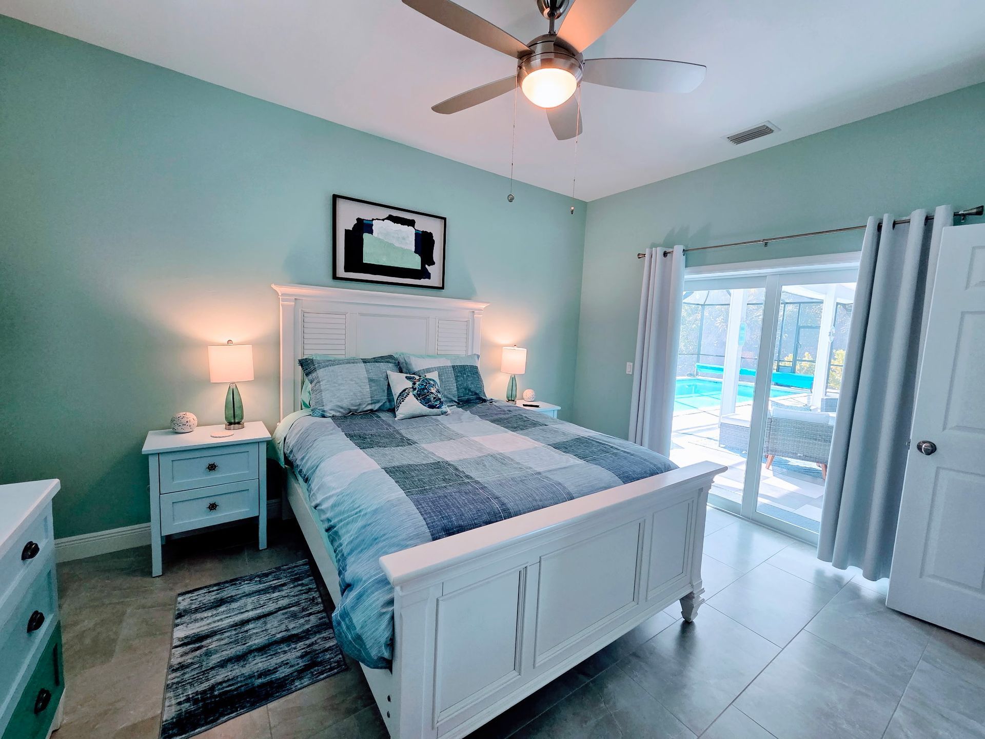 A bedroom with pale green walls, a white bed, bedside tables, a ceiling fan, and sliding doors leading to a pool area.