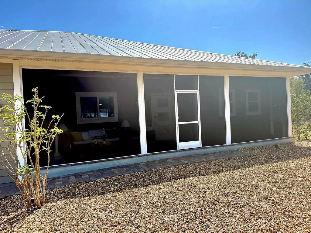 A house with a large, screened-in porch, white support pillars, and a metal roof, viewed from a gravel yard.
