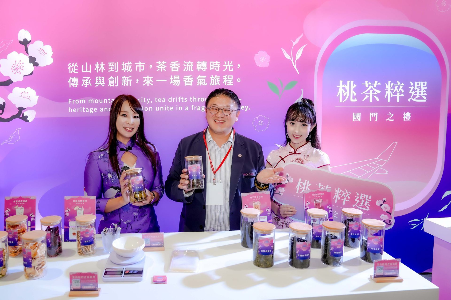 Three people at a tea promotion: holding tea jars, smiling, with a banner and product display.