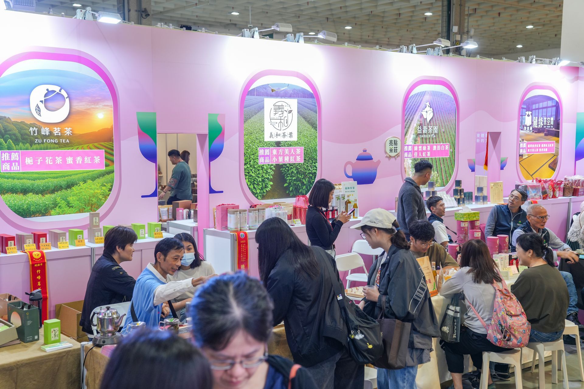 People browsing tea products at a trade show booth, pink walls with scenic views, tea packaging on display.