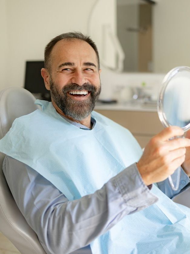 A man is sitting in a dental chair looking at his teeth in a mirror.