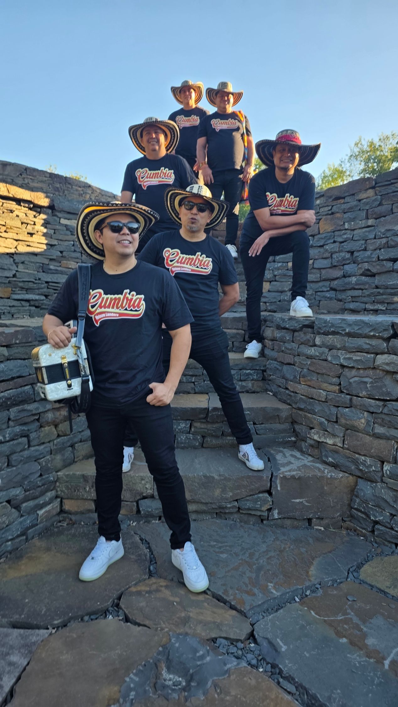 Grupo de seis personas con camisas negras y sombreros posando en escalones de piedra.
