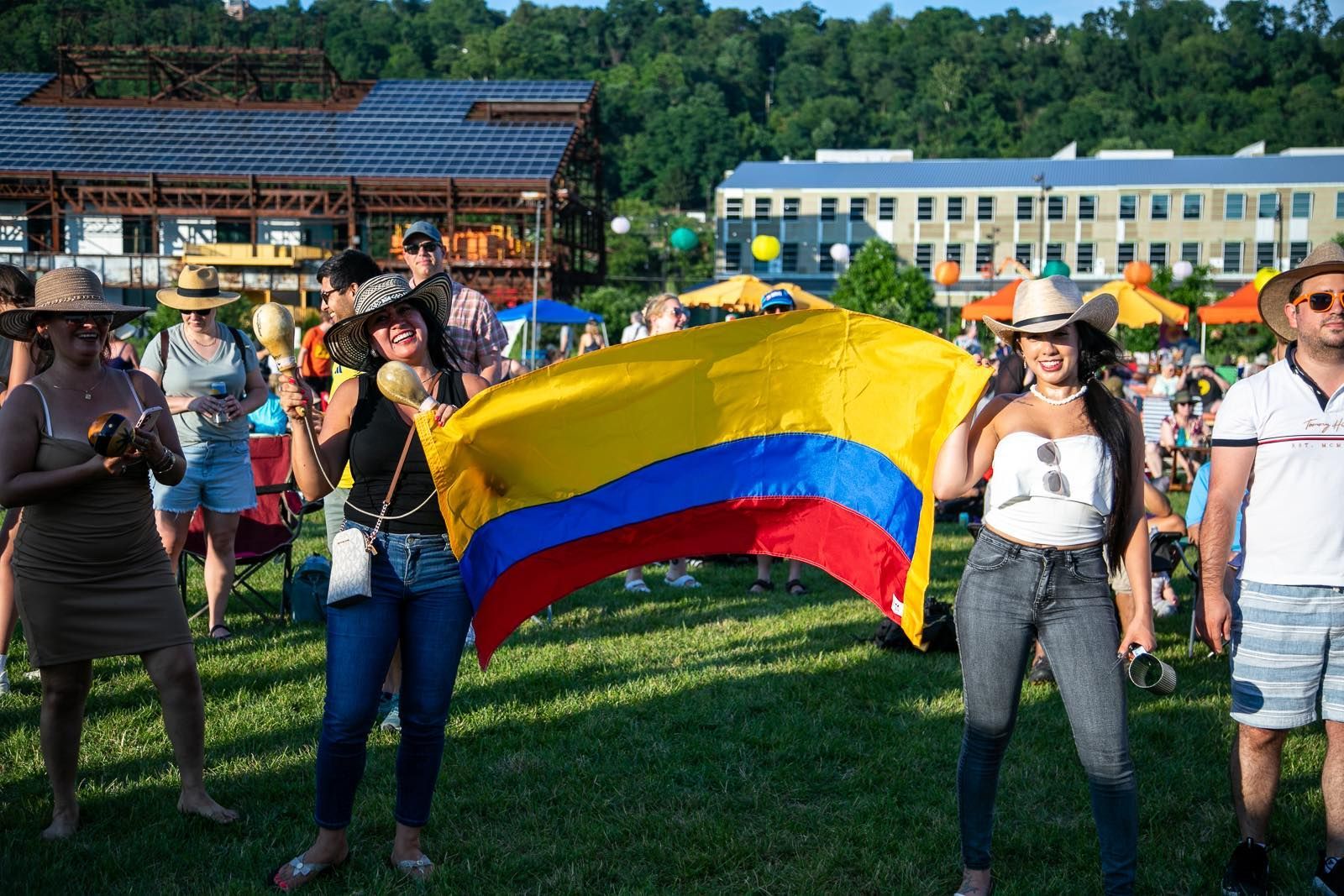 Gente celebrando con una bandera colombiana. Al aire libre, soleado, con maracas y sonriendo.