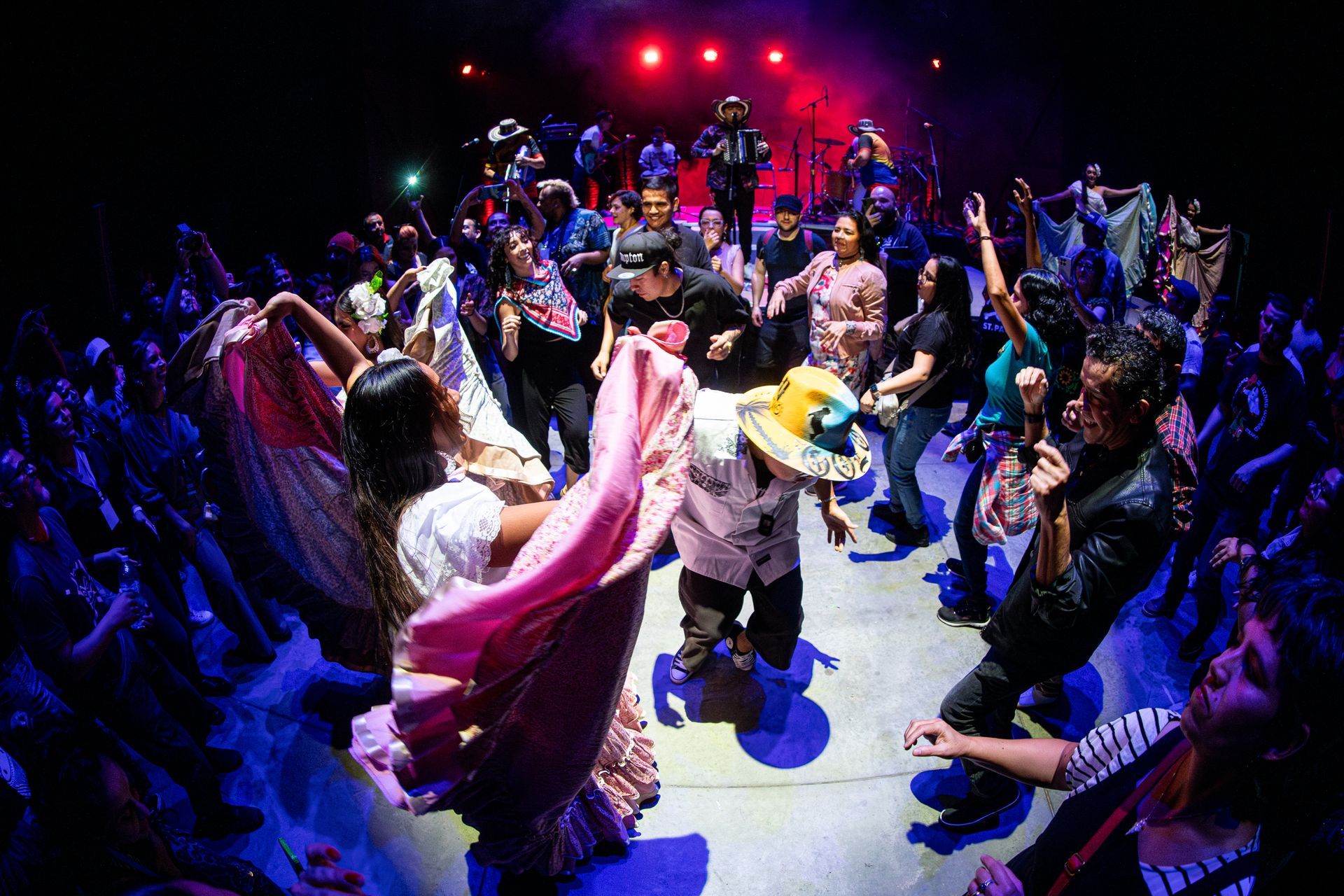 Un grupo animado bailando en un concierto con una banda en el escenario; gente con ropa colorida, con los brazos en alto