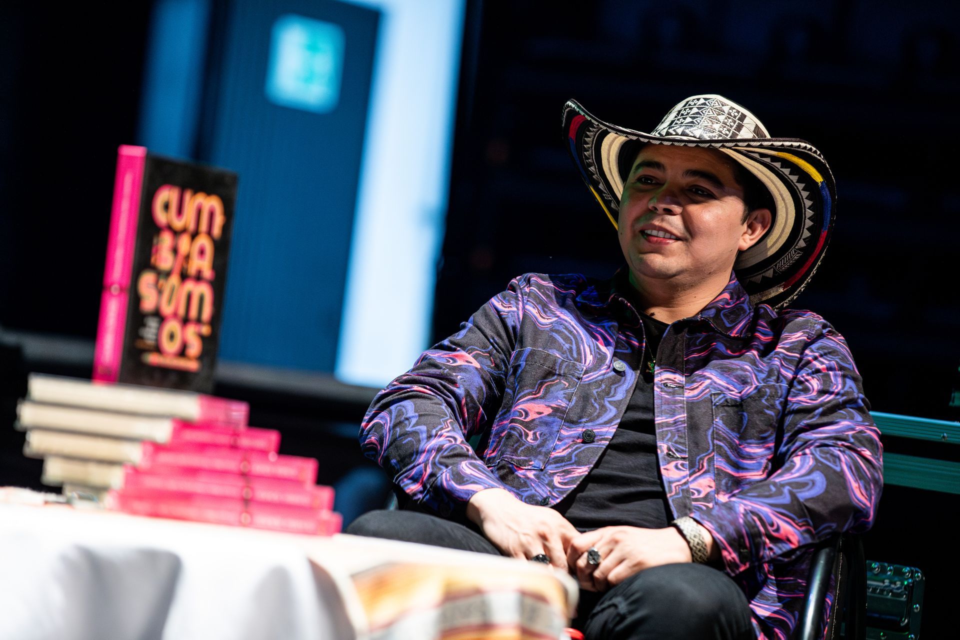 Hombre con chaqueta estampada y sombrero sentado a la mesa con libros; escenario.