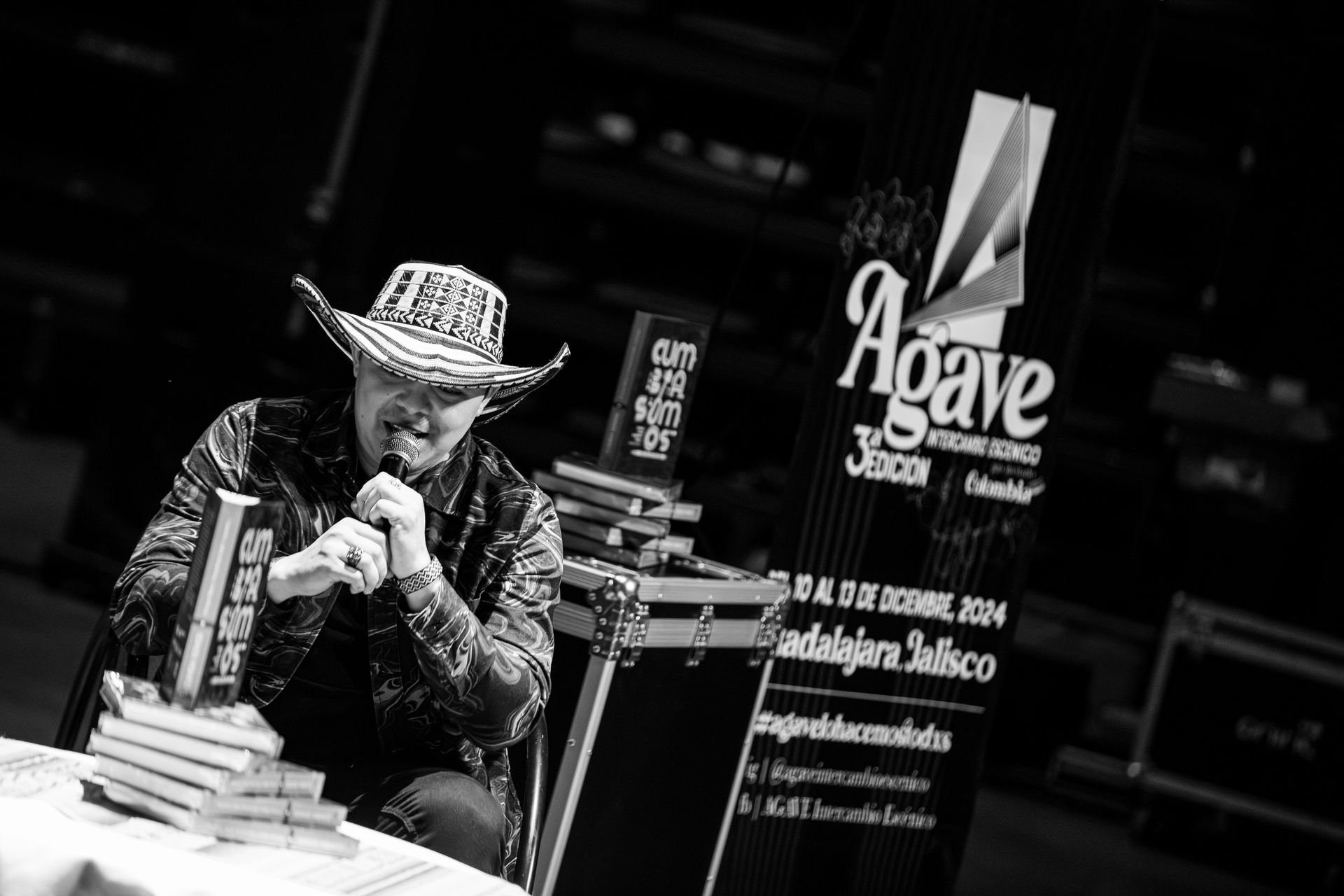 Hombre firmando libros en la Feria del Libro de Guadalajara, con sombrero y camisa estampada. Foto en blanco y negro.