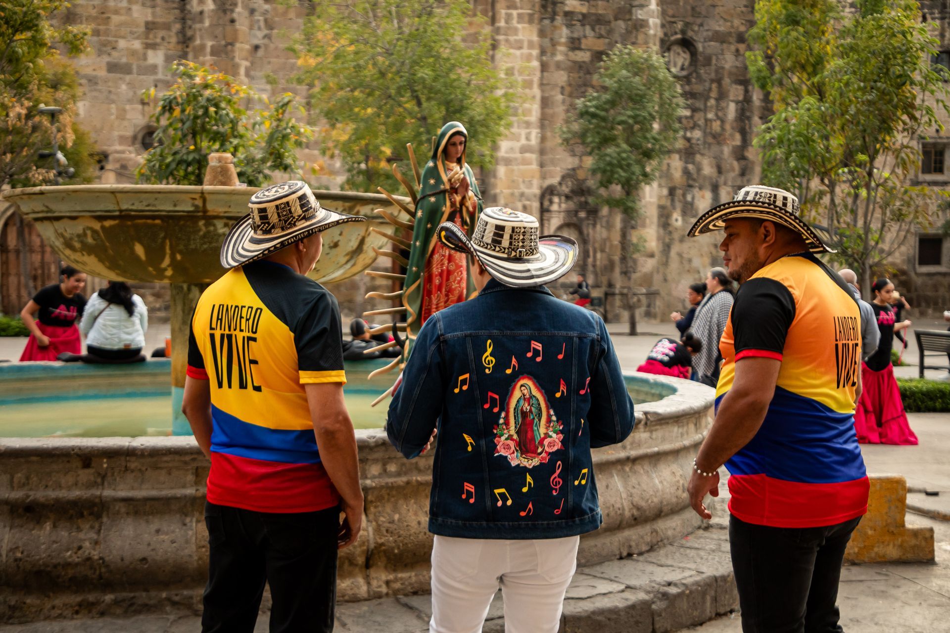 Tres hombres con camisas y sombreros coloridos están cerca de una fuente, con una estatua de la Virgen María al fondo.