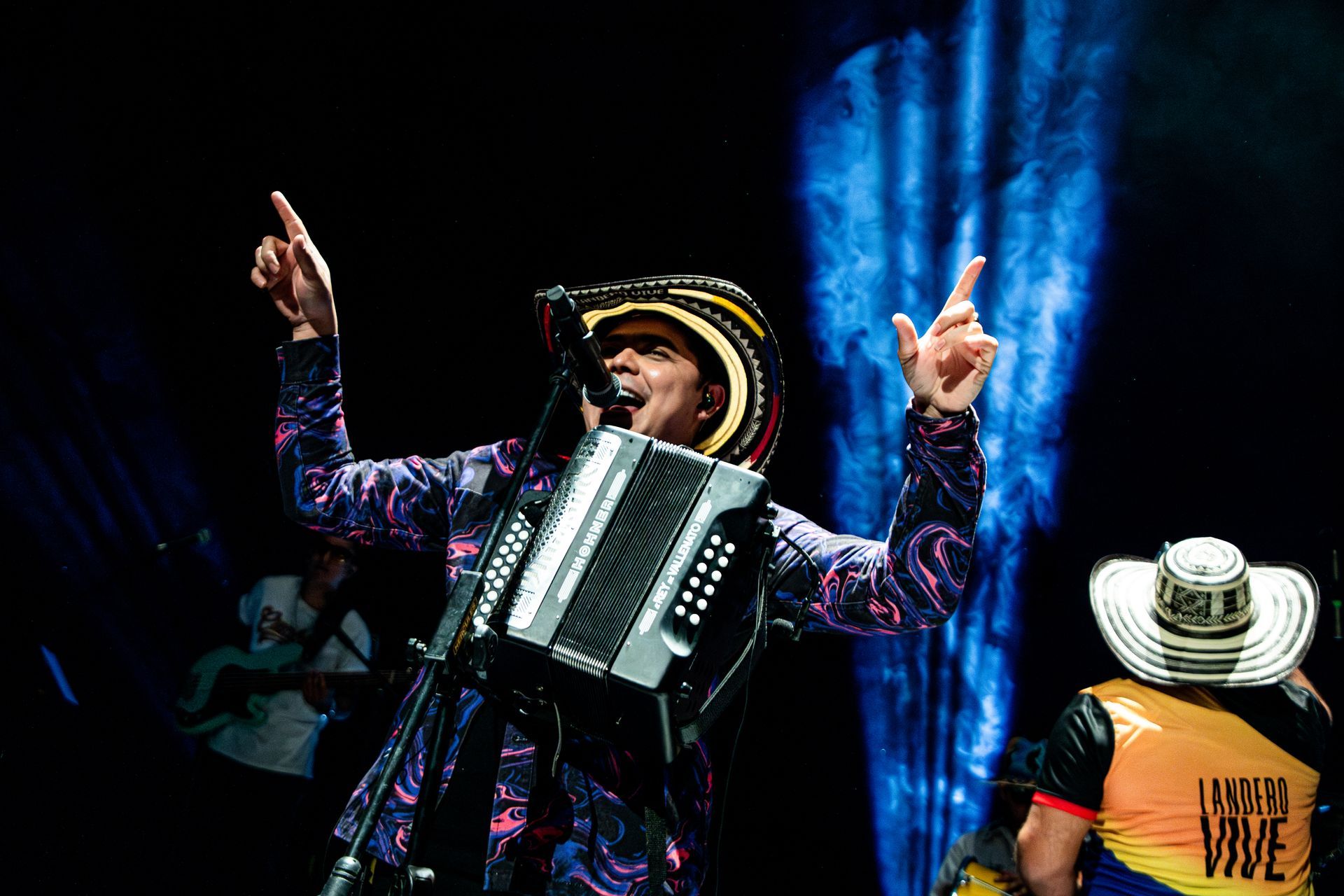 Hombre tocando el acordeón, con sombrero, apuntando hacia el escenario, con otro músico al fondo.