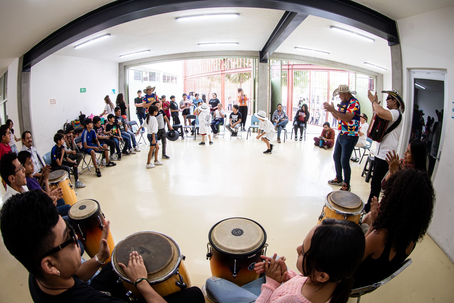 Personas en círculo tocando instrumentos, cantando y bailando en una habitación luminosa y abierta.