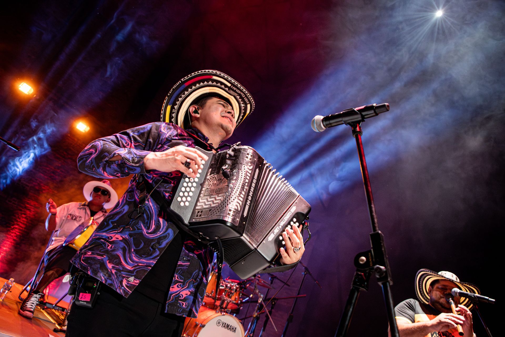 Hombre tocando el acordeón en el escenario, vistiendo un sombrero colorido y una camisa estampada
