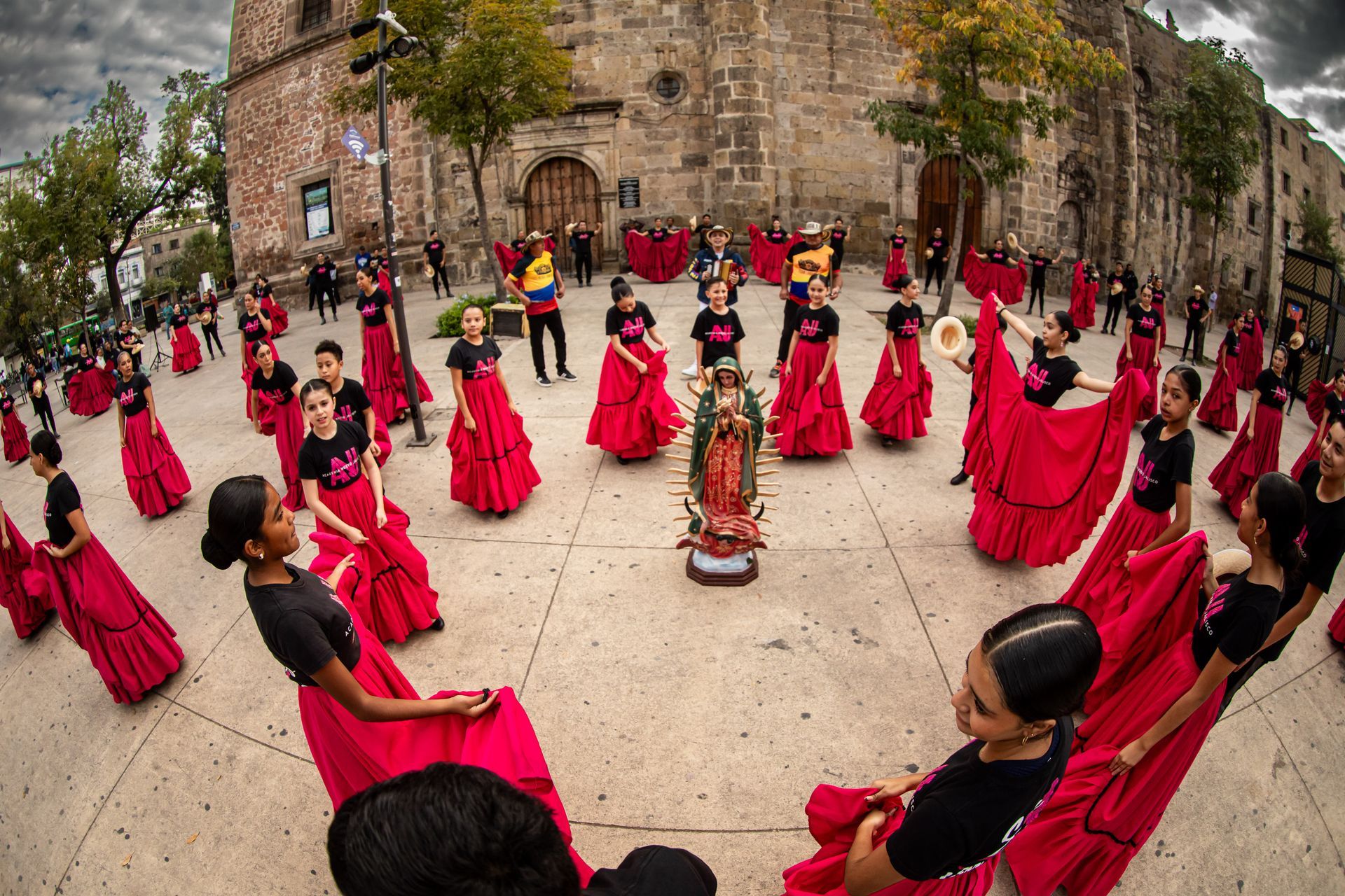 Bailarines con faldas rojas y camisas negras rodean una estatua de la Virgen María en una plaza.