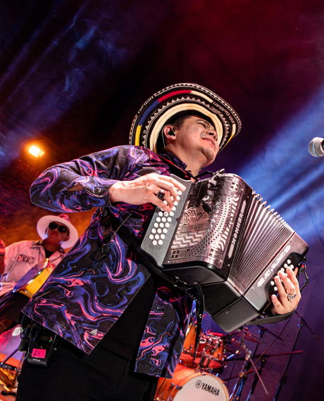 Un hombre con una camisa colorida toca el acordeón en el escenario, usando un sombrero.